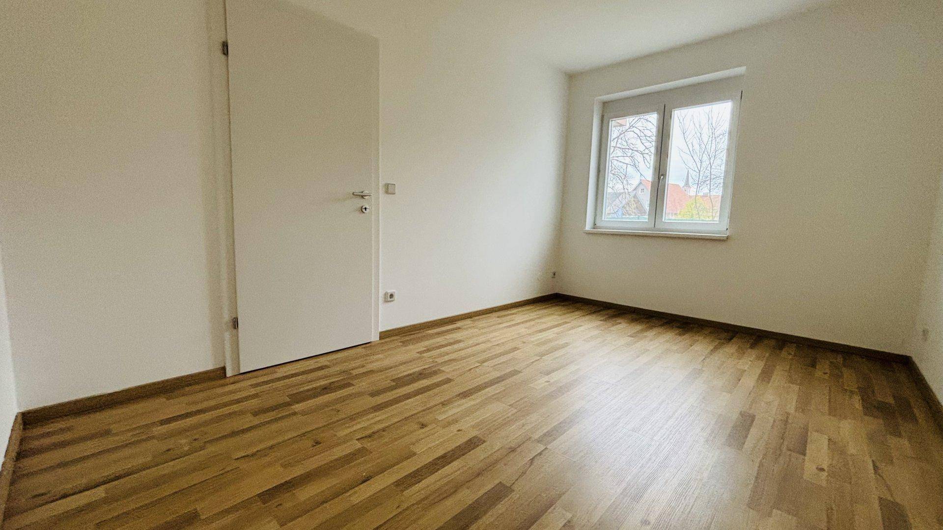 Helles Schlafzimmer mit Parkettboden und großem Fenster für viel Tageslicht.