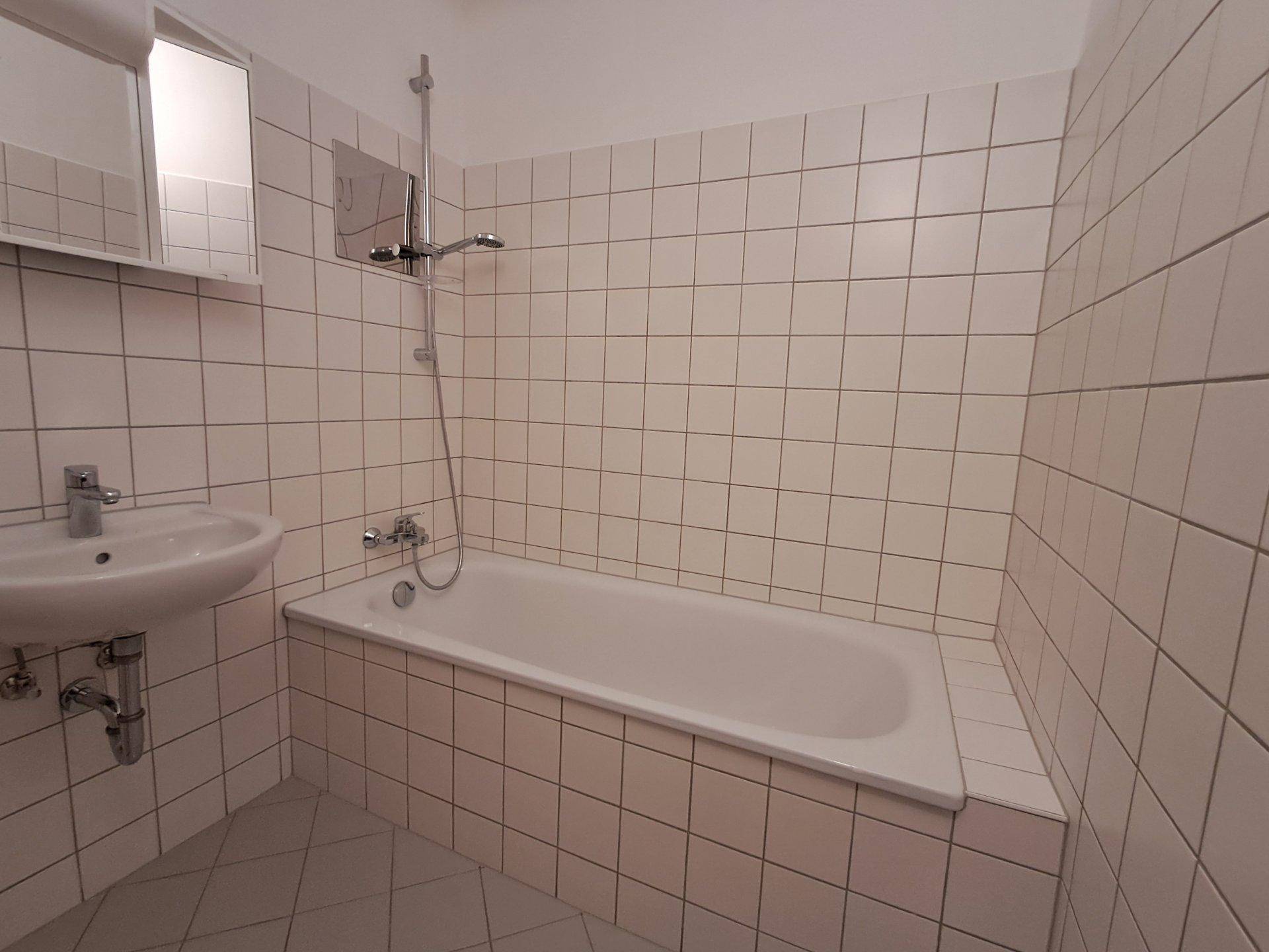 Badezimmer mit weißen Fliesen, Badewanne mit Duschfunktion und einem einzelnen Waschbecken.