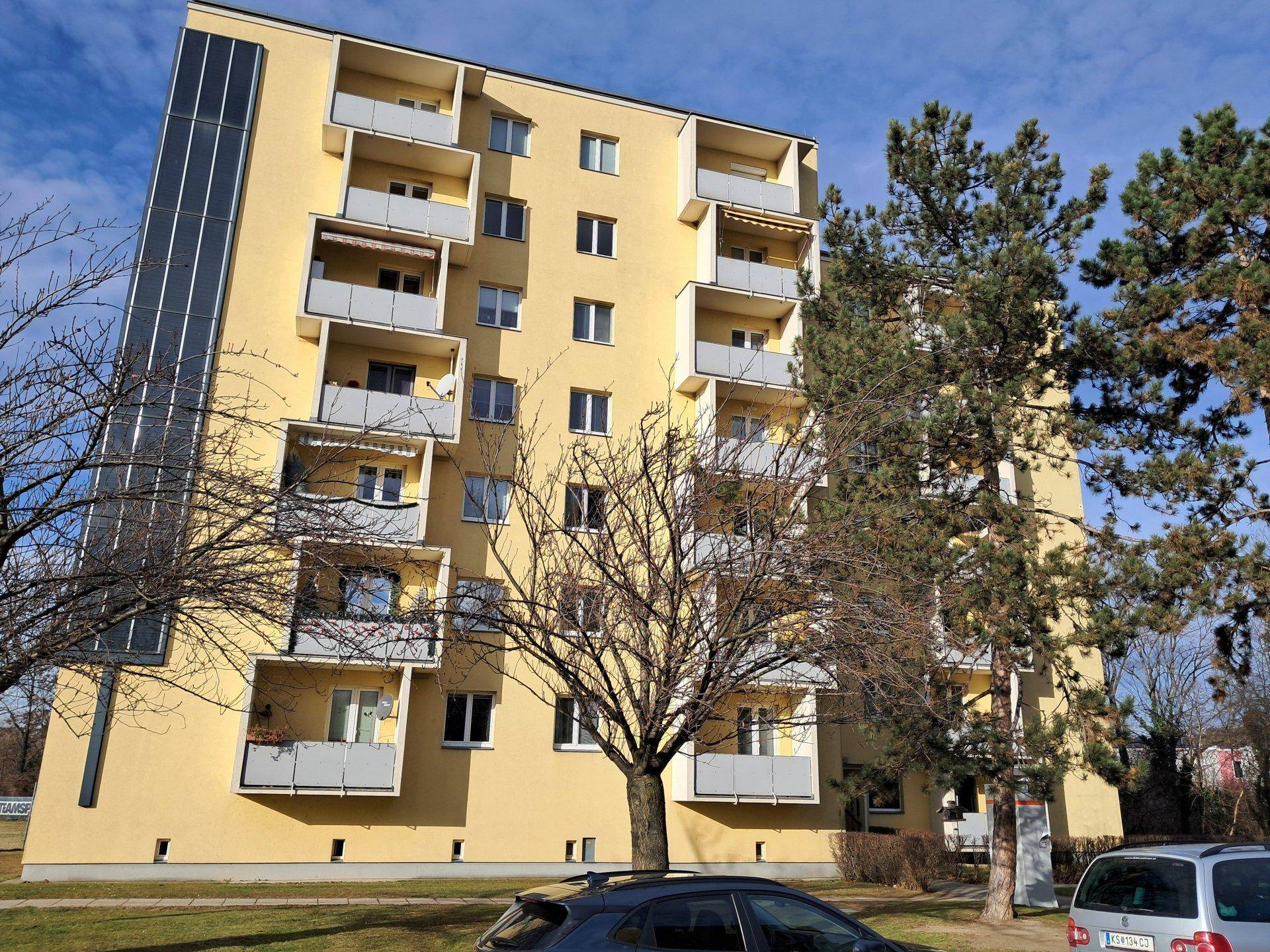 Mehrstöckiges gelbes Wohnhaus mit Balkonen und Solarpaneelen, umgeben von Bäumen im Winter.