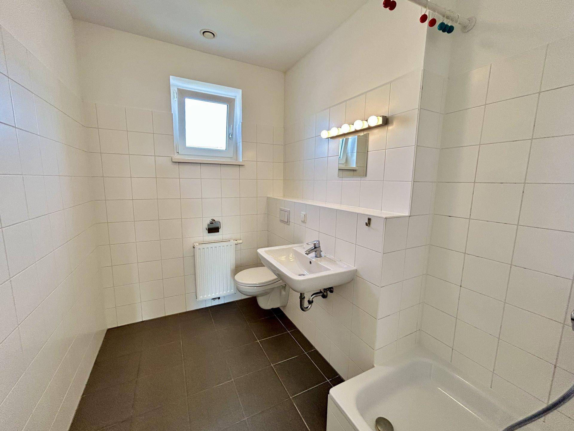 Helles Badezimmer mit weißen Fliesen, Fenster, Waschbecken, WC und einer kleinen Badewanne.