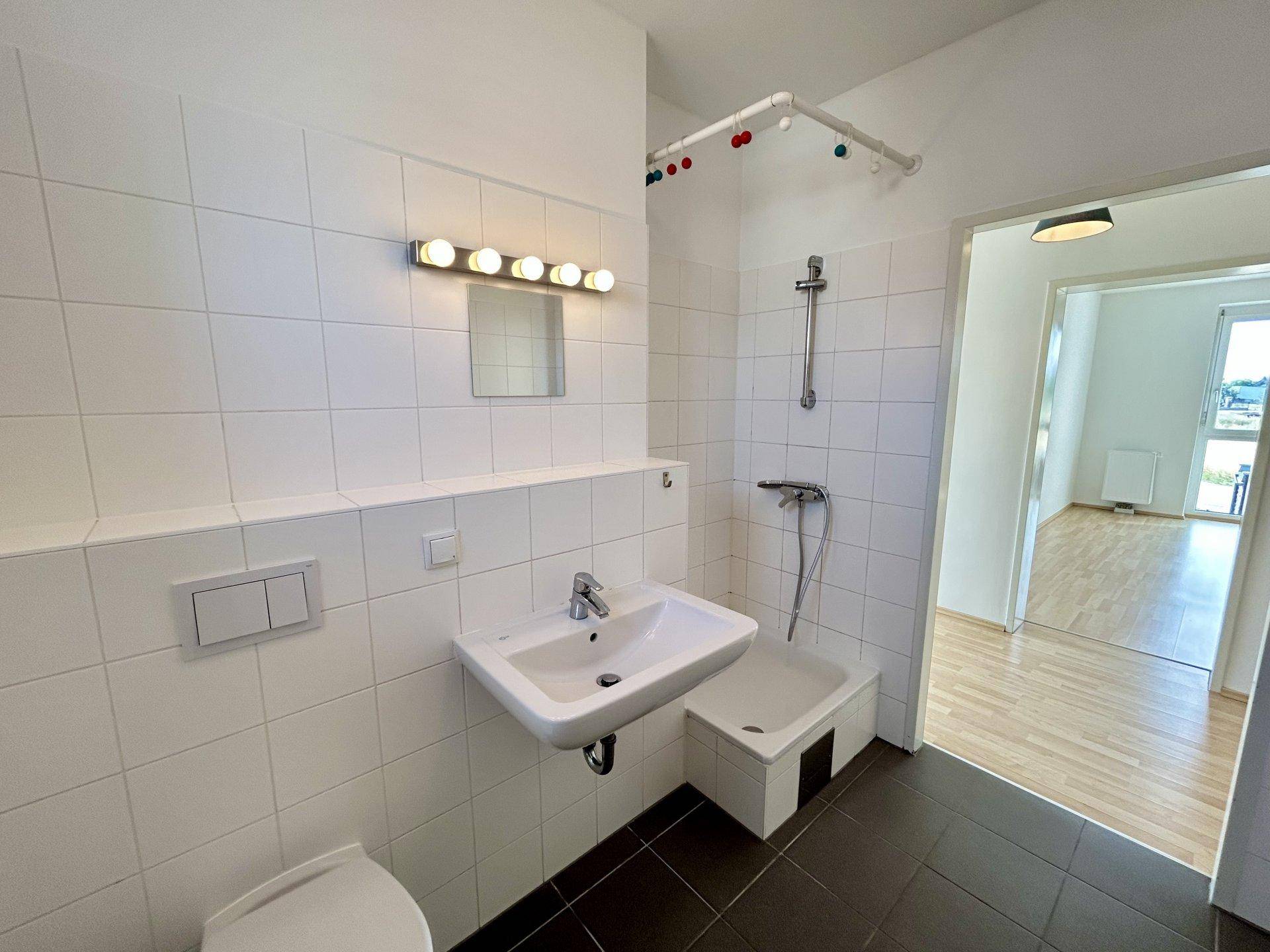Funktionales Badezimmer mit weißen Wandfliesen, dunklem Boden, Waschbecken, WC und Duschbereich.