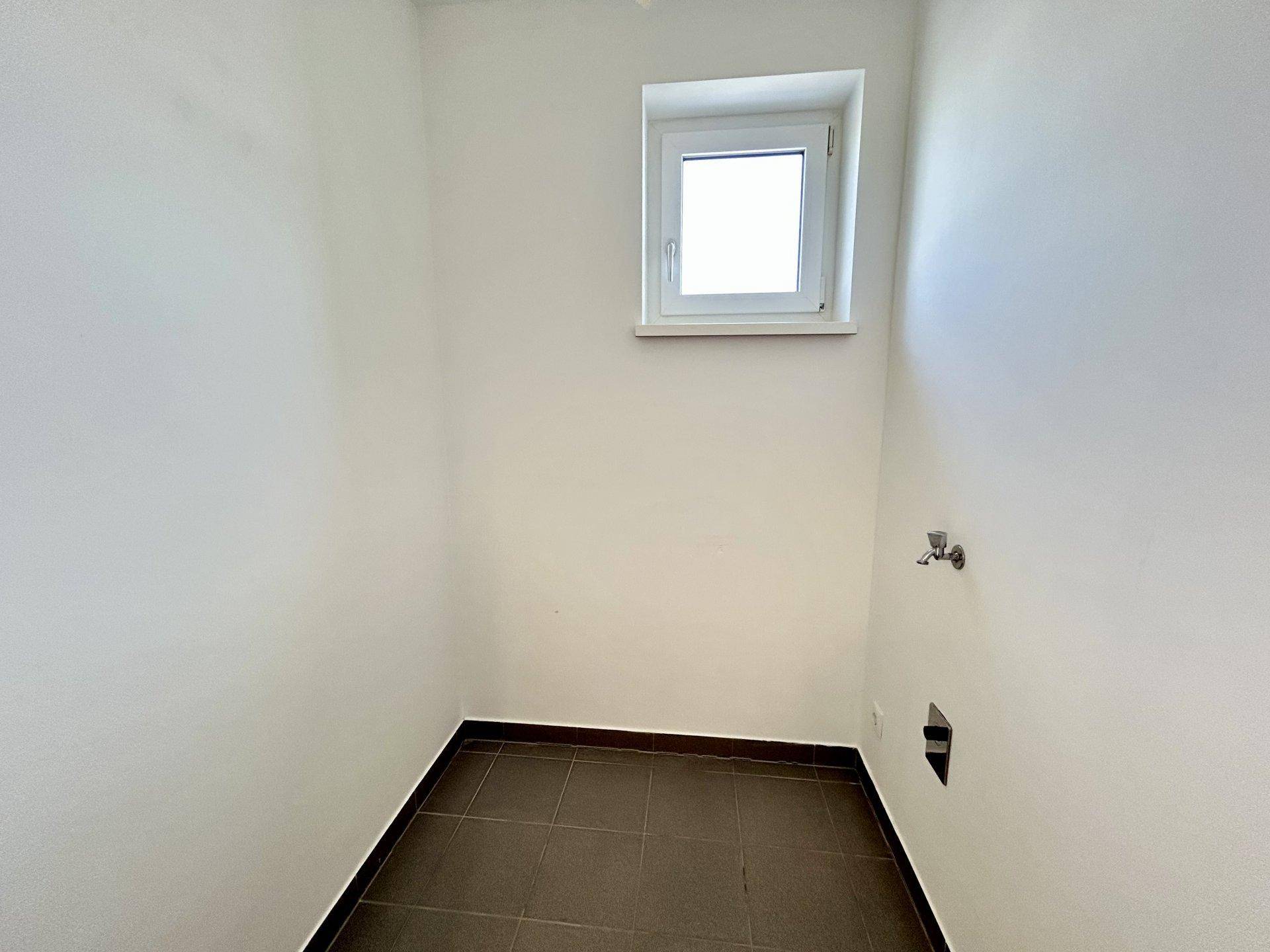 Kleiner Hauswirtschaftsraum mit dunklen Fliesen, einem Fenster und Anschlüssen für Geräte.