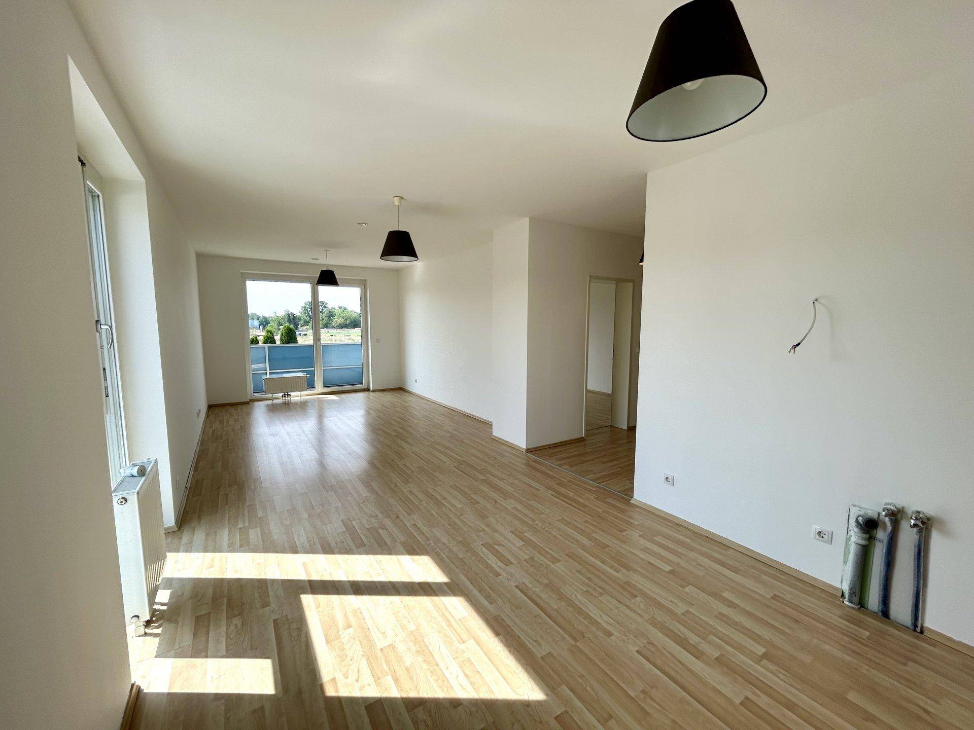 Geräumiger, heller Wohnbereich mit Laminatboden, großen Fenstern und Zugang zu einem Balkon.