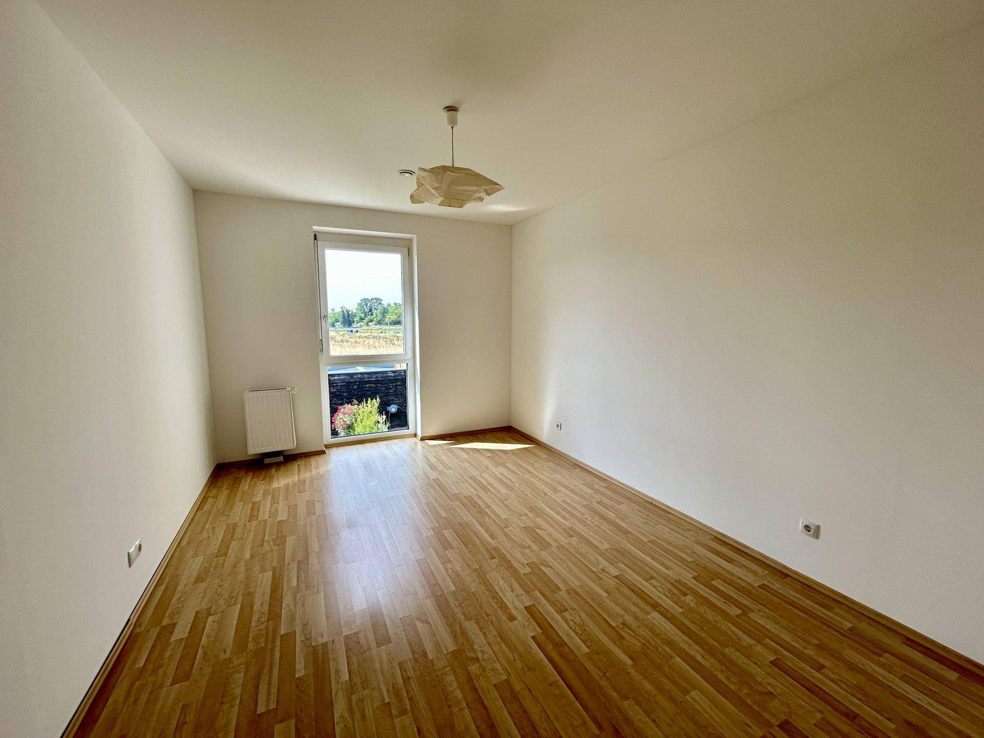Leeres Zimmer mit Laminatboden, weißen Wänden und Zugang zu einem Außenbereich durch eine große Tür.