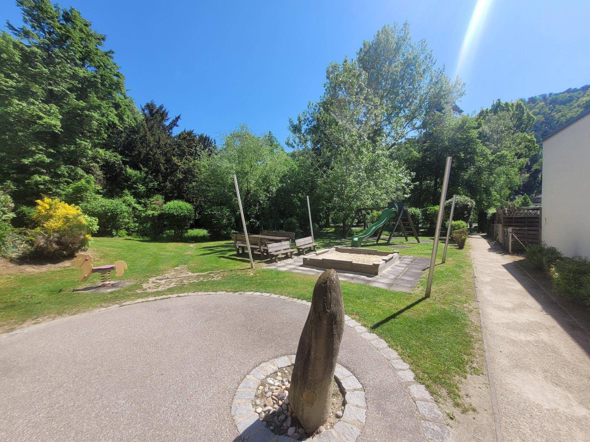 Spielplatz mit Sandkasten, Rutsche und Schaukelpferd, umgeben von Bäumen und Grünflächen.