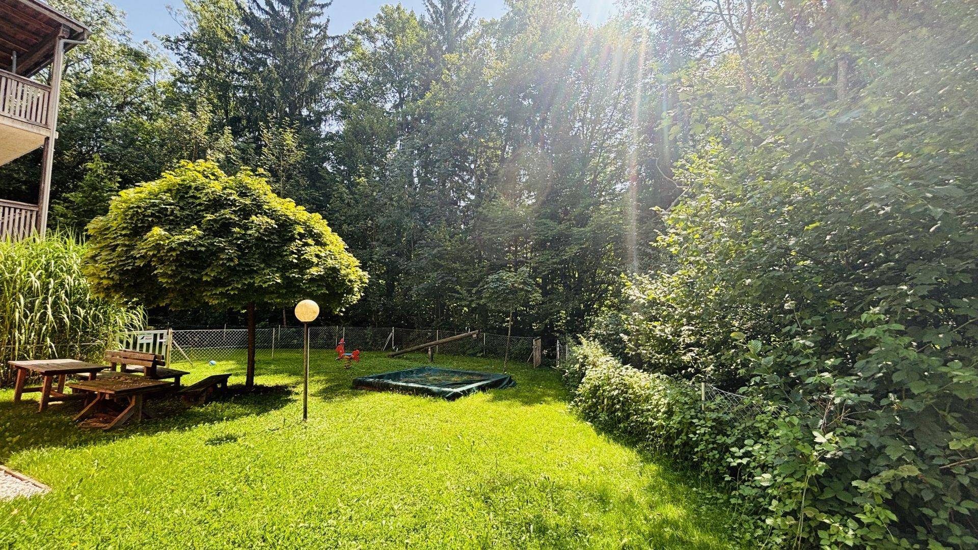 Sonniger Gemeinschaftsgarten mit Spielplatz, Sandkasten und Sitzgelegenheiten, umgeben von Bäumen.