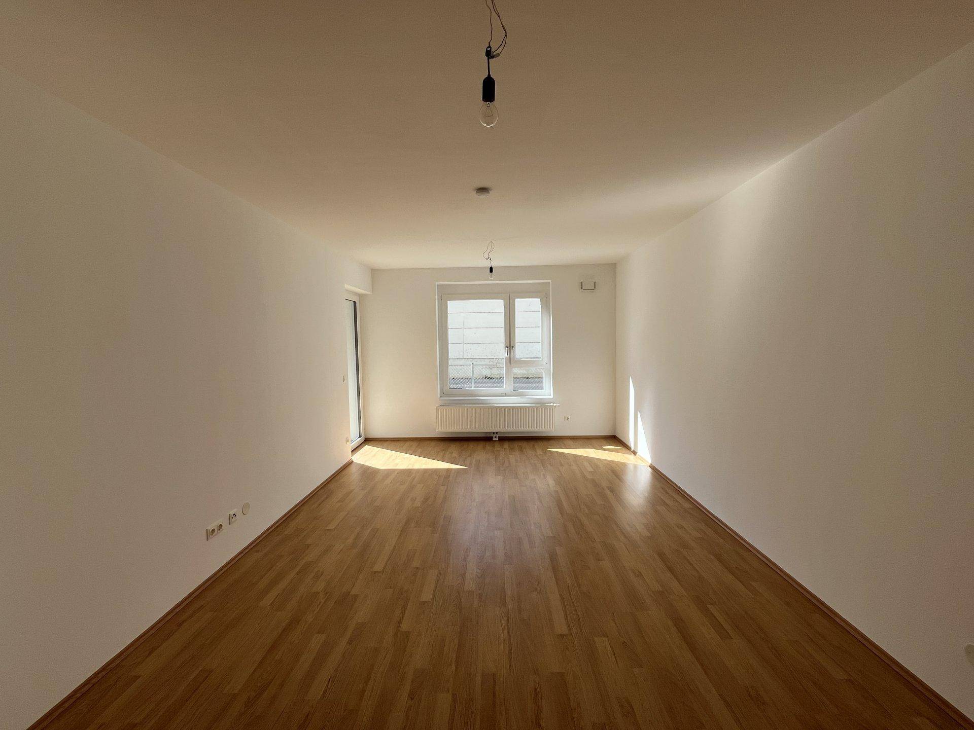 Leeres, helles Wohnzimmer mit Holzparkettboden und einem Fenster, das viel Tageslicht hereinlässt.