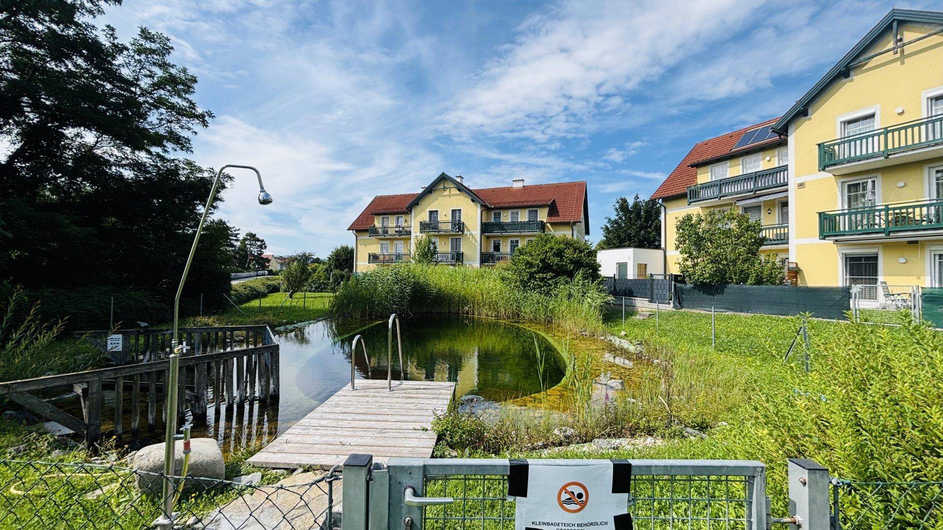 Natürlicher Badeteich mit Holzsteg und Außendusche vor einem gelben Wohngebäude.