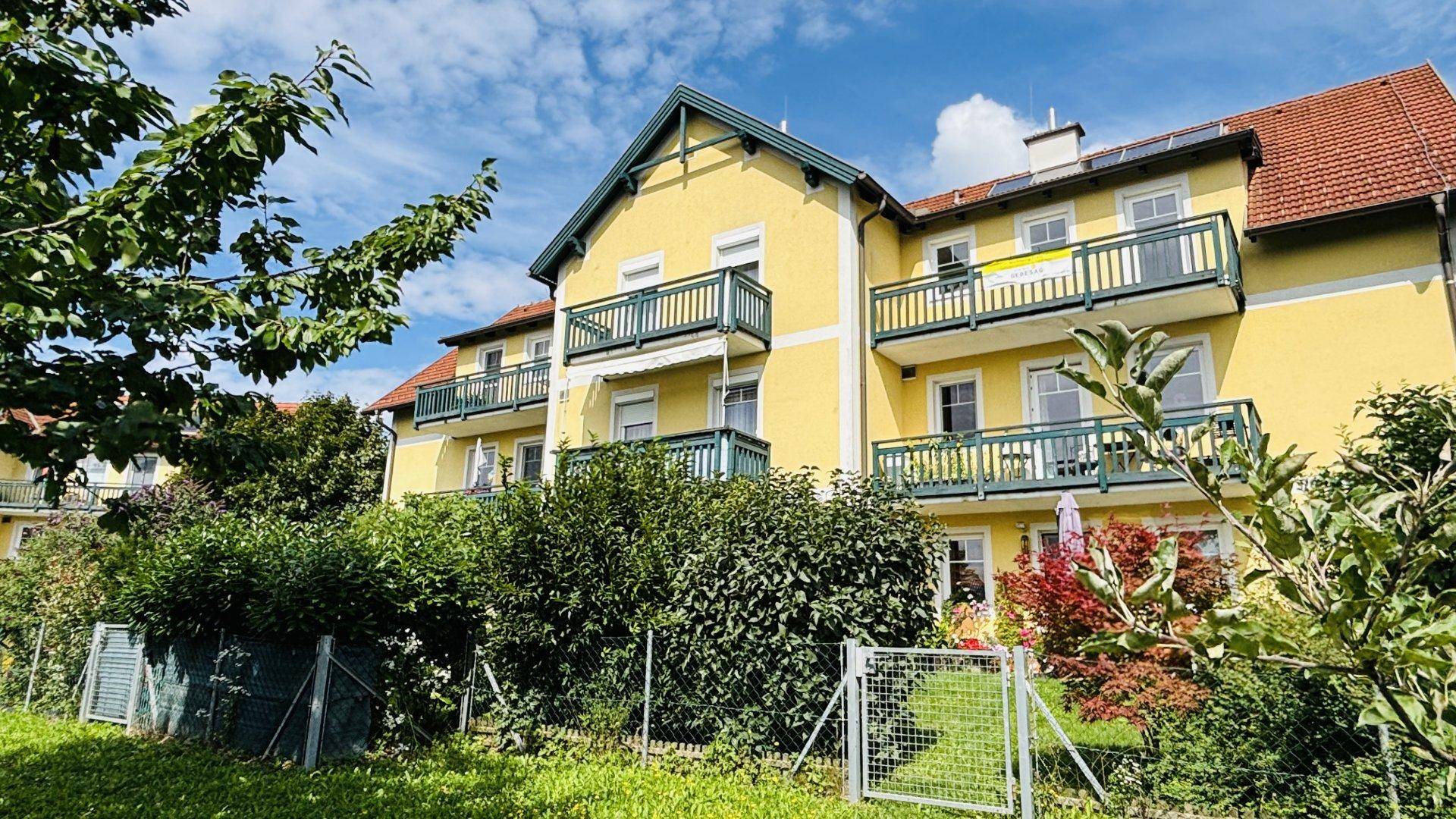 Gelbes Wohnhaus mit grünen Balkonen und roten Dächern, umgeben von gepflegten Hecken und Bäumen.