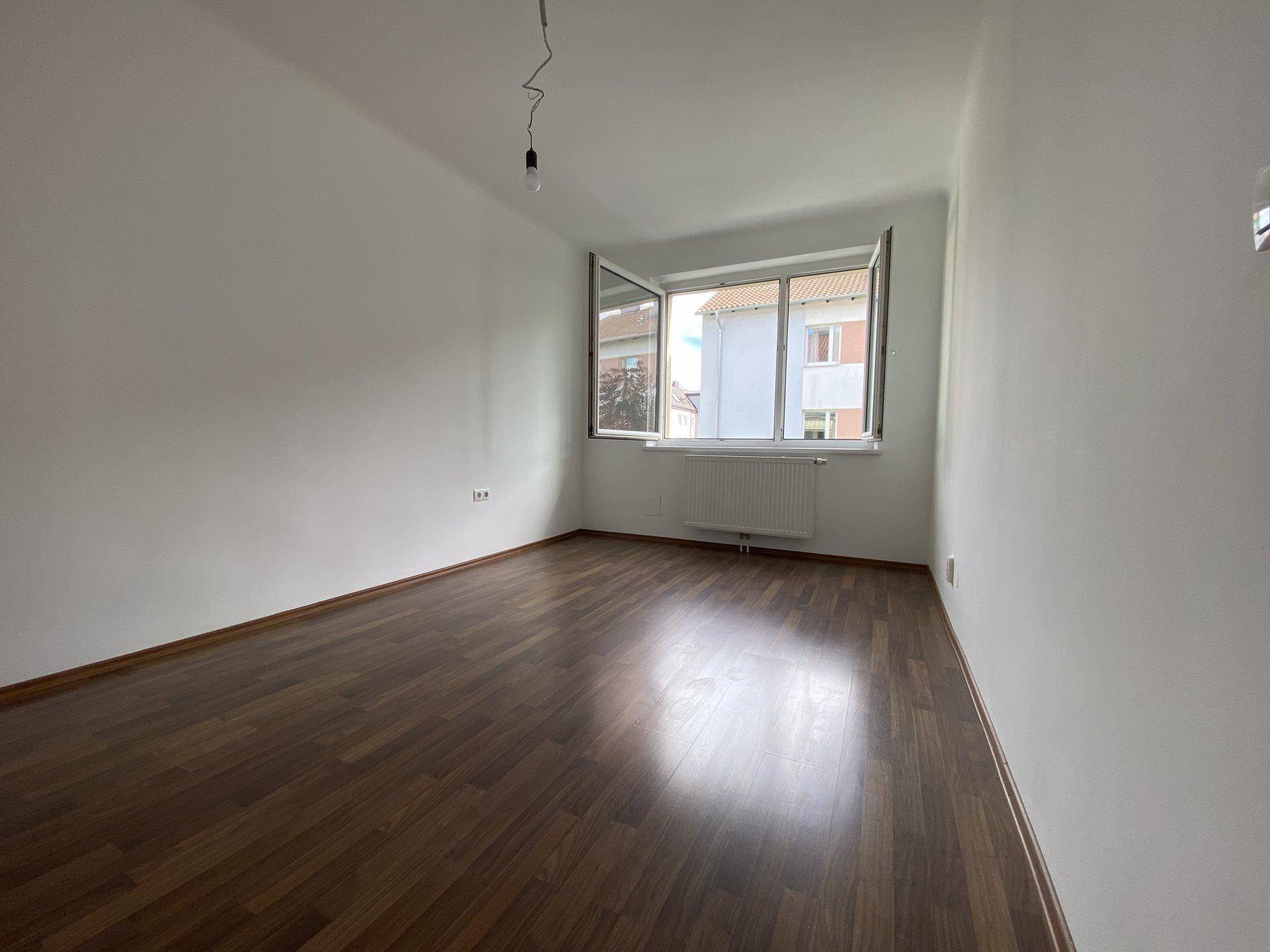 Helles, leeres Wohnzimmer mit dunklem Laminatboden und großem Fenster.