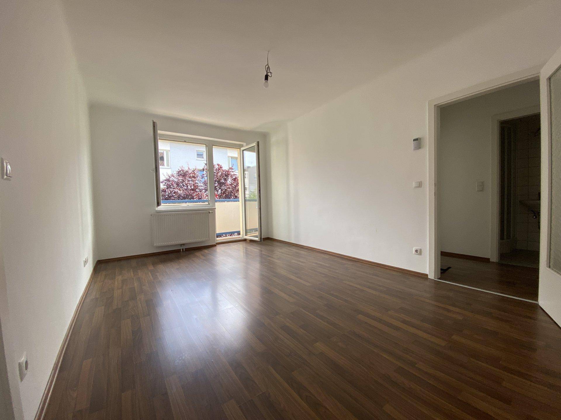 Geräumiges Wohnzimmer mit Laminatboden, Balkonzugang und Blick ins Grüne.