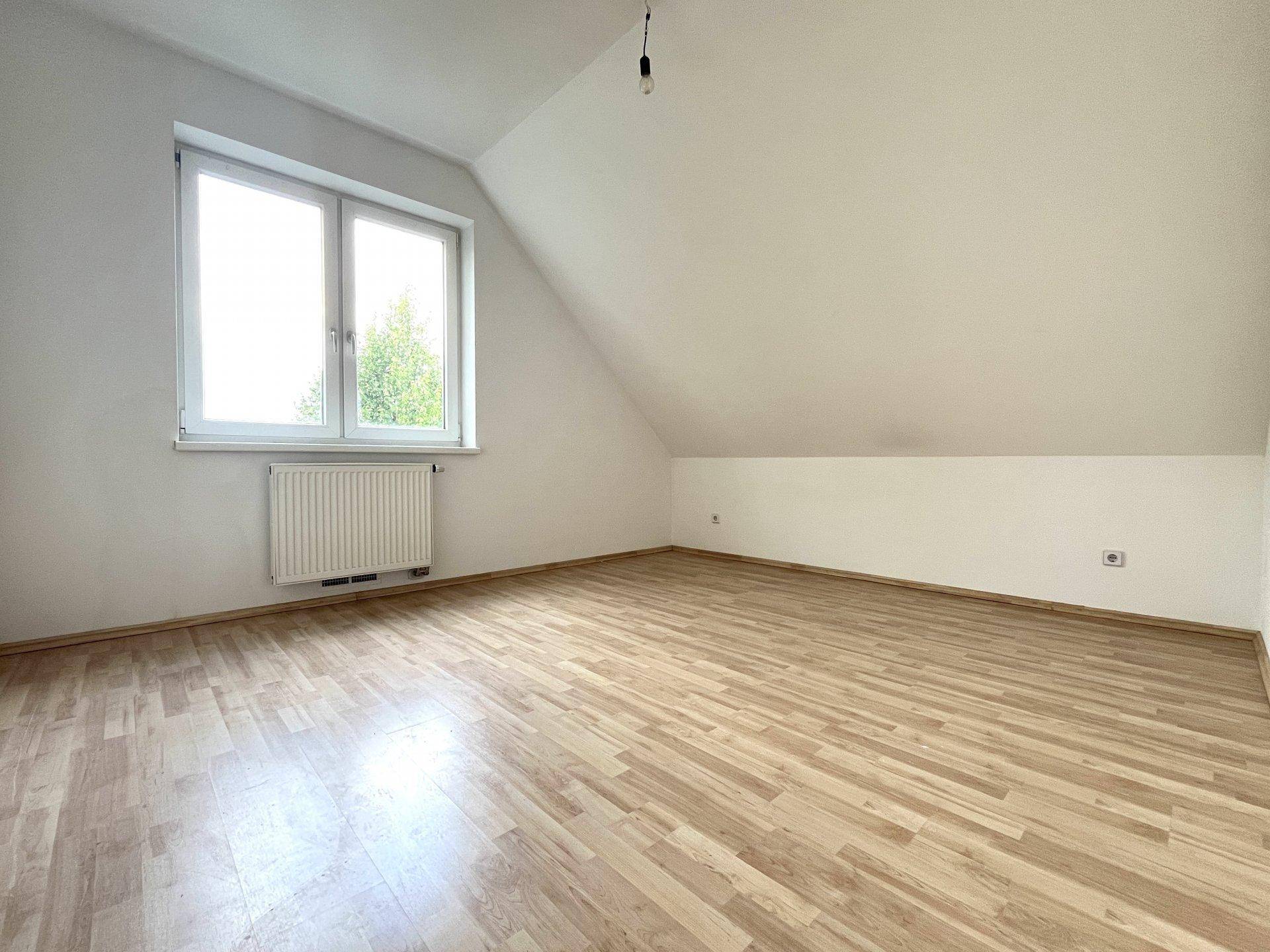 Geräumiges Schlafzimmer mit schrägen Wänden, hellem Laminatboden und einem großen Fenster.