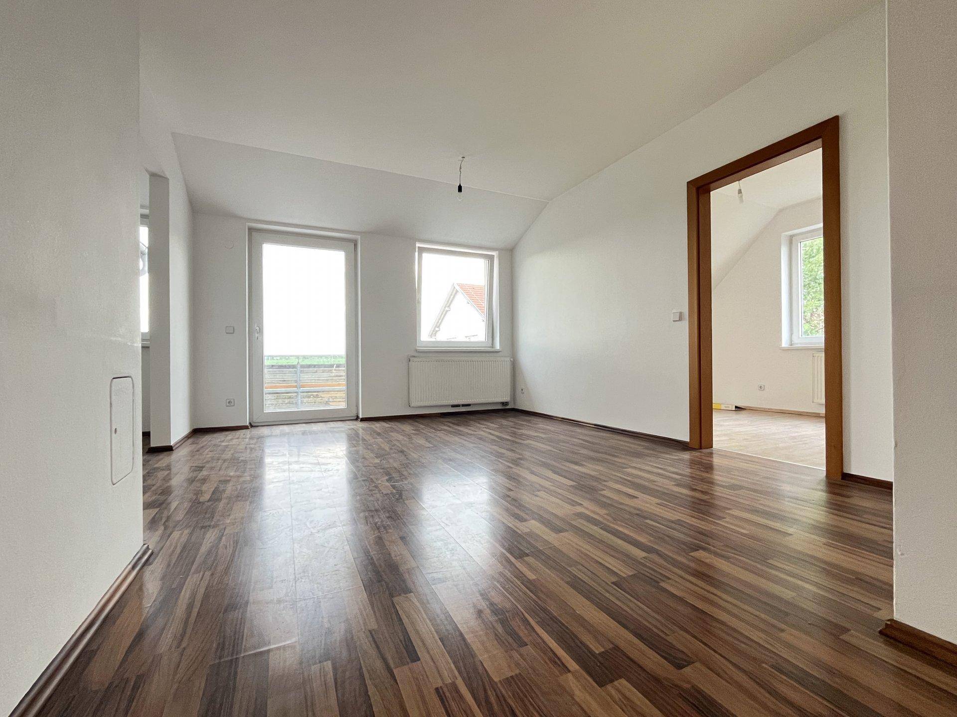 Geräumiger, leerer Raum mit dunklem Laminatboden, weiß gestrichenen Wänden und Zugang zum Balkon.