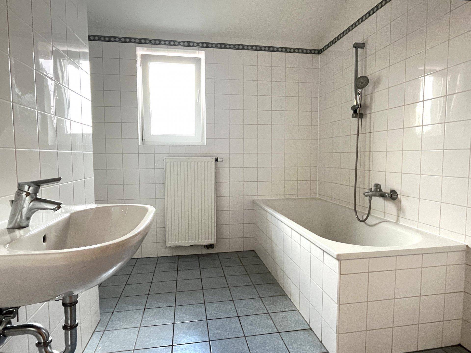 Gefliestes Badezimmer mit Badewanne, Waschbecken, Fenster und blauen Bodenfliesen.