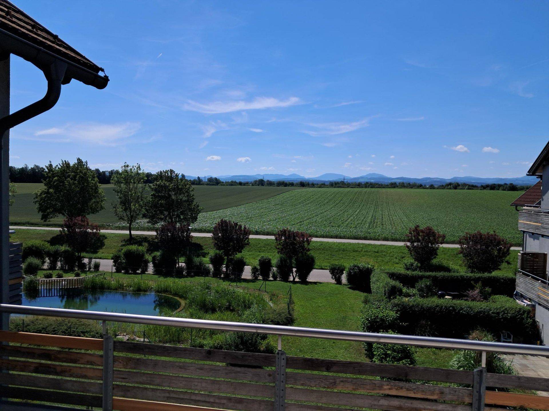 Sonniger Balkonblick auf einen Teich, weite Felder und ferne Berge unter blauem Himmel.