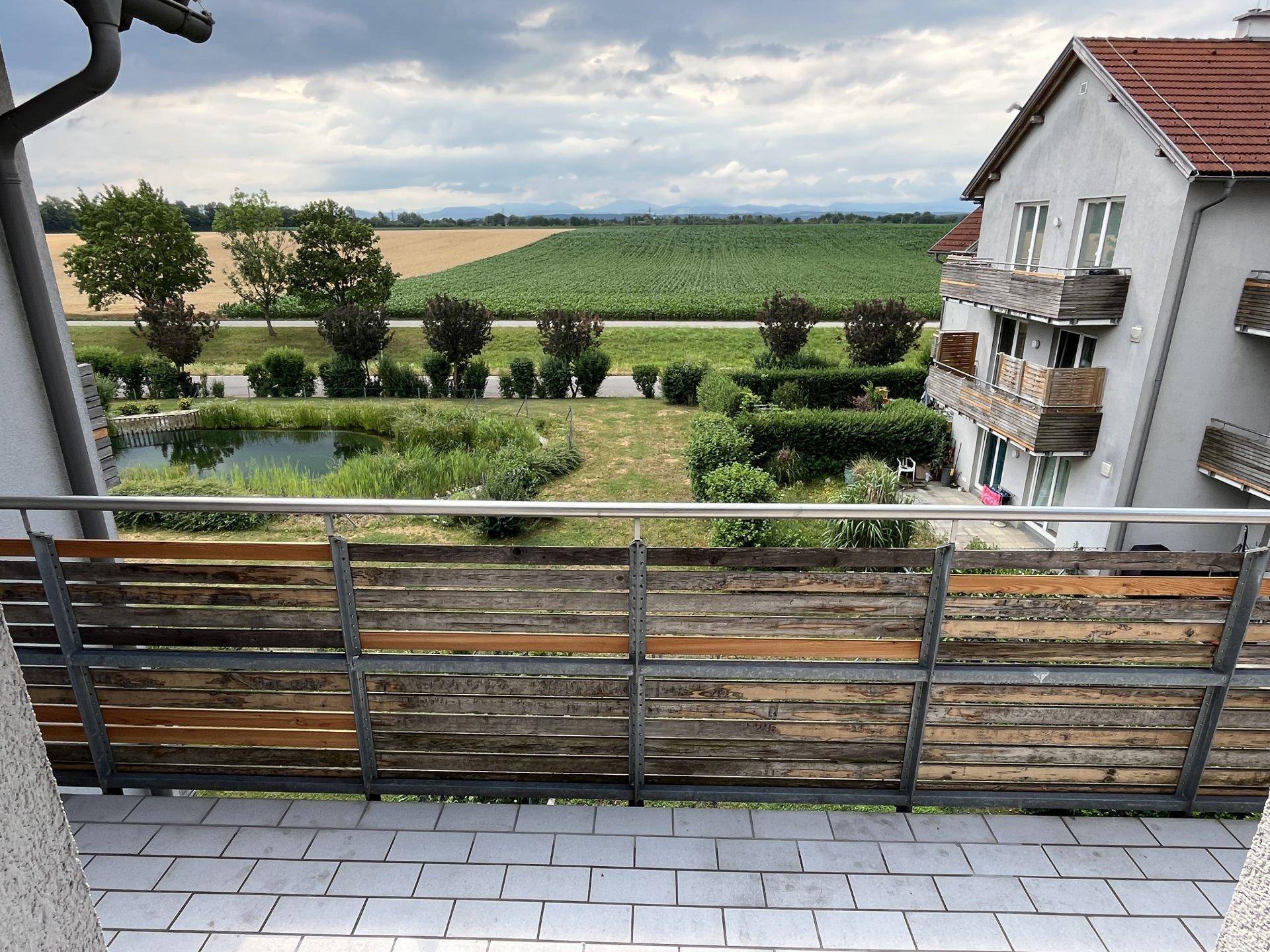 Balkonblick auf einen Teich, grüne Felder und eine Bergkette unter bewölktem Himmel.
