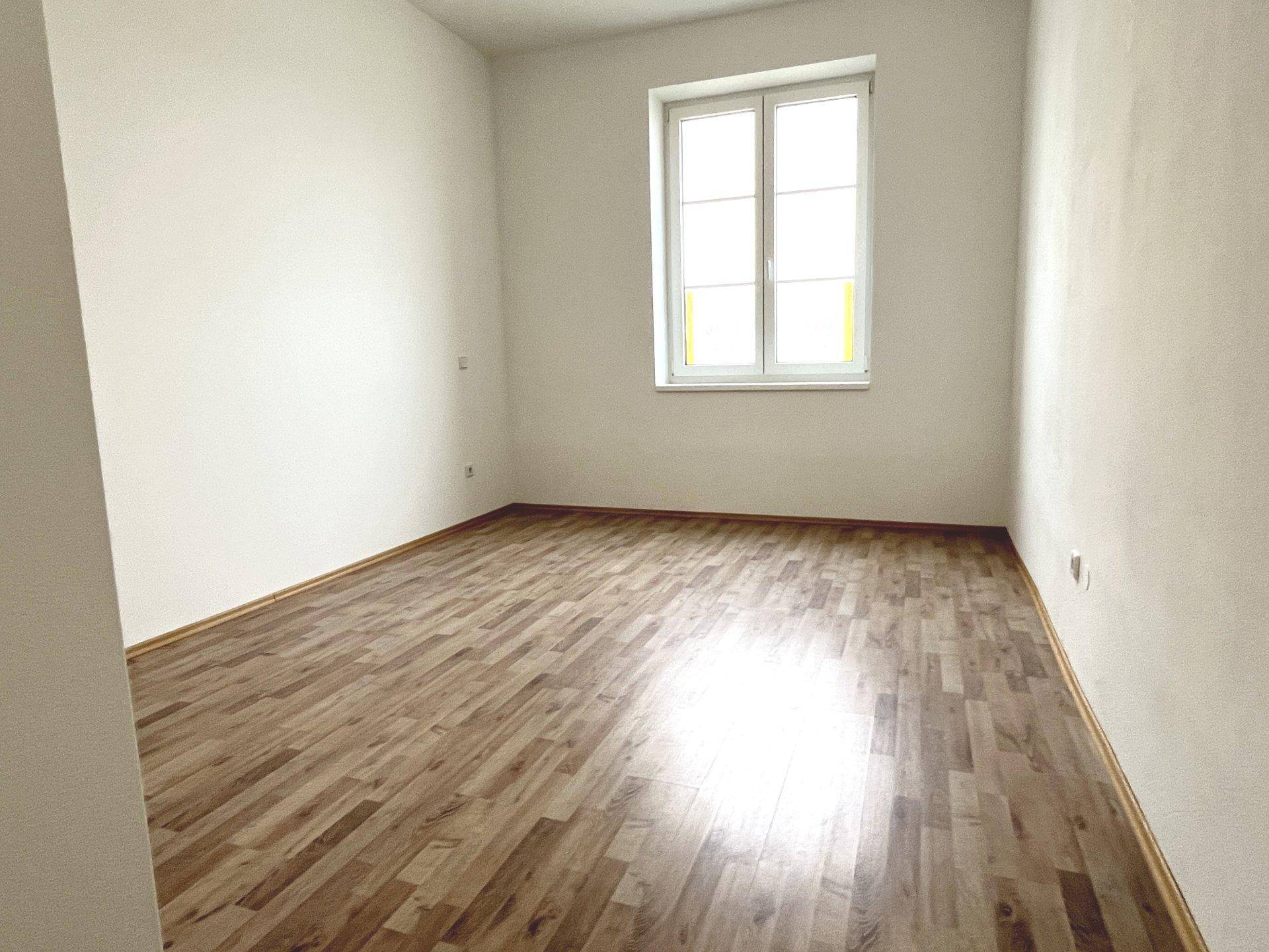 Geräumiger, heller Raum mit Laminatboden und einem großen Fenster, das viel Tageslicht hereinlässt.