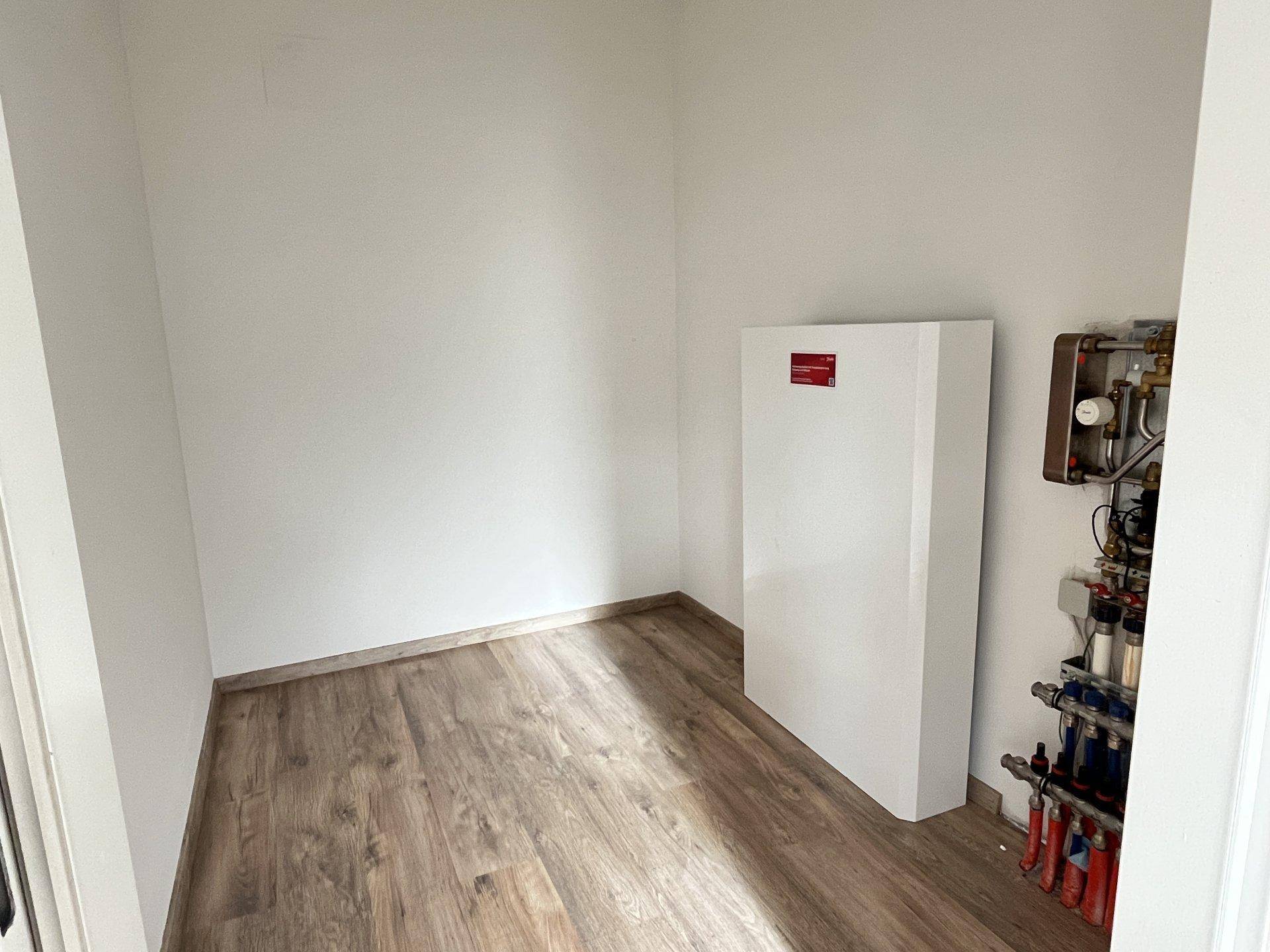 Kleiner Hauswirtschaftsraum mit Wärmepumpe und Heizungsinstallationen auf Laminatboden.
