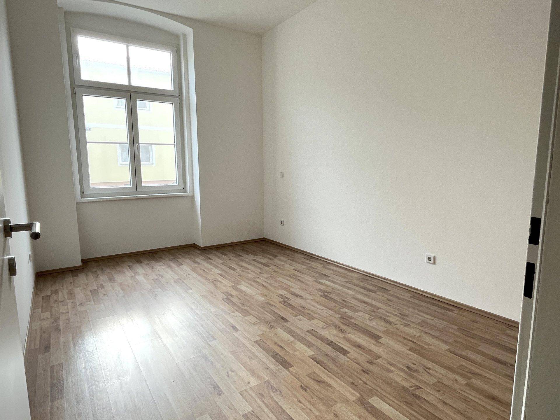 Heller, leerer Raum mit Laminatboden und einem großen Fenster, das auf die Straße blickt.