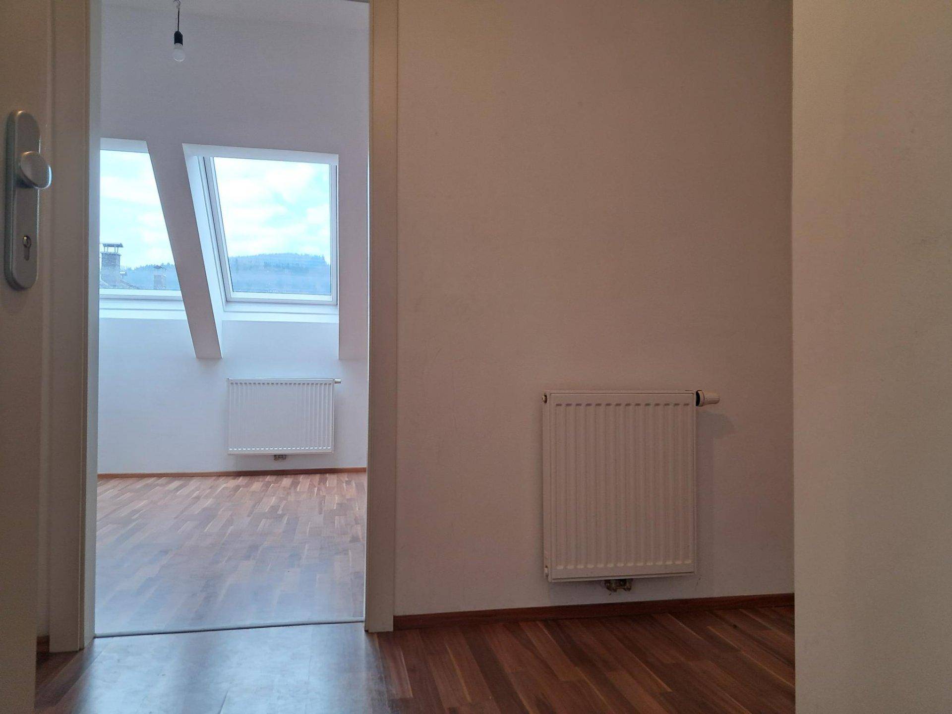 Heller Raum im Dachgeschoss mit Dachfenstern und Holzfußboden, gesehen durch eine Türöffnung.