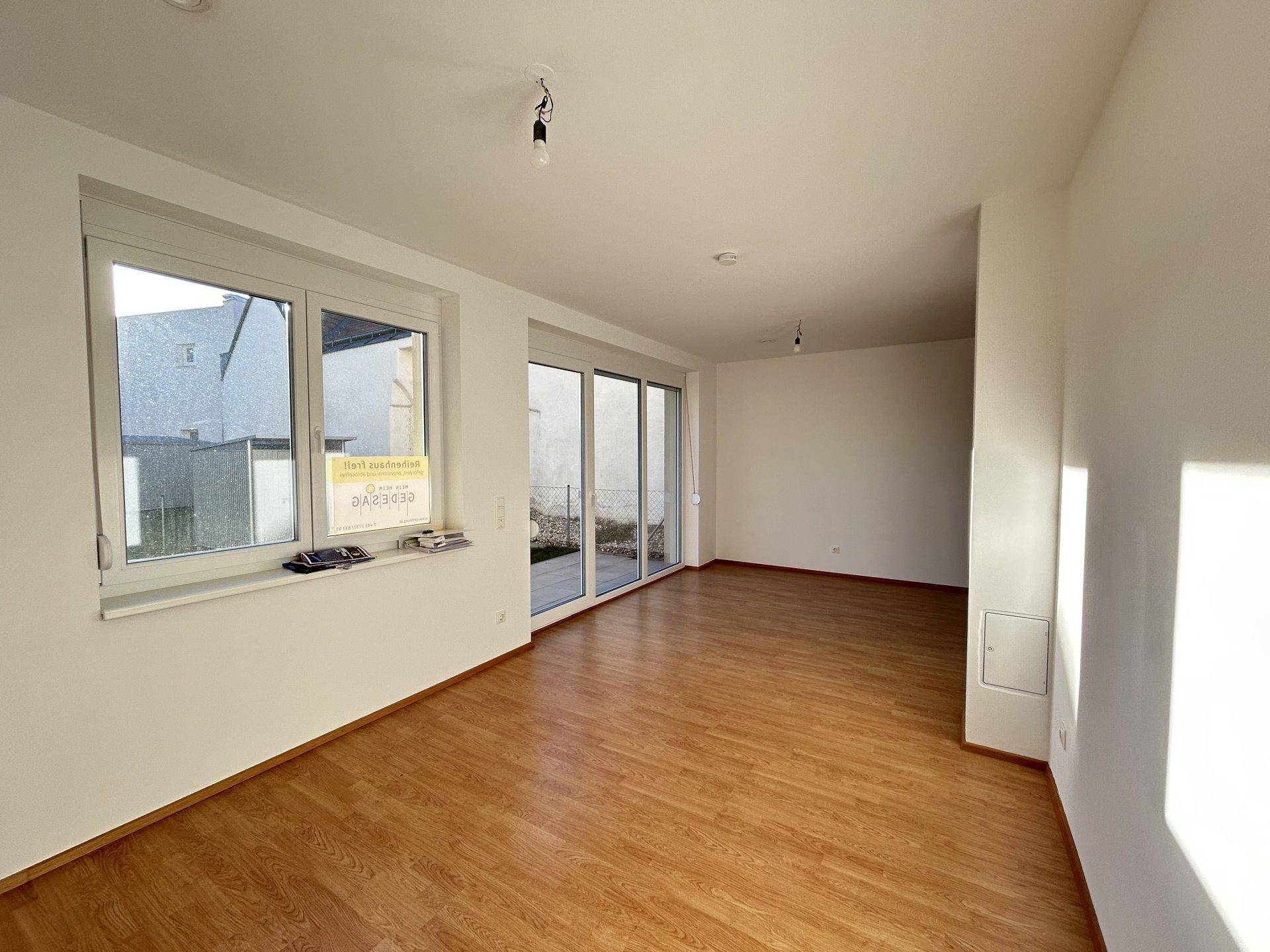 Geräumiges, leeres Wohnzimmer mit Laminatboden und großen Fenstern, die viel Licht hereinlassen.