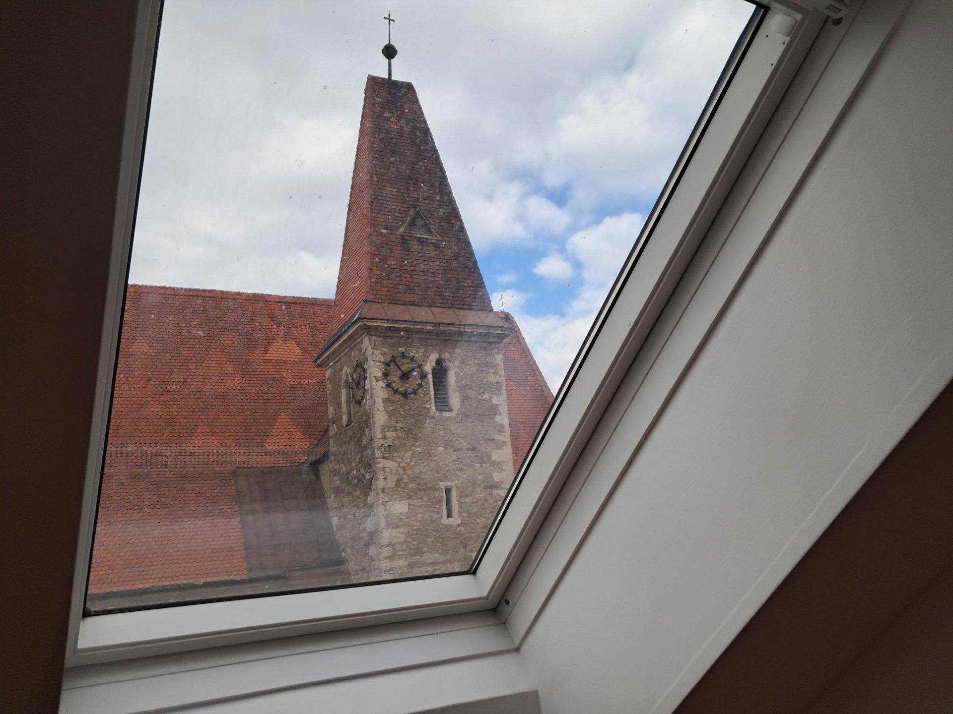 Blick aus einem Dachfenster auf einen Kirchturm mit Uhr und umliegende Dächer.