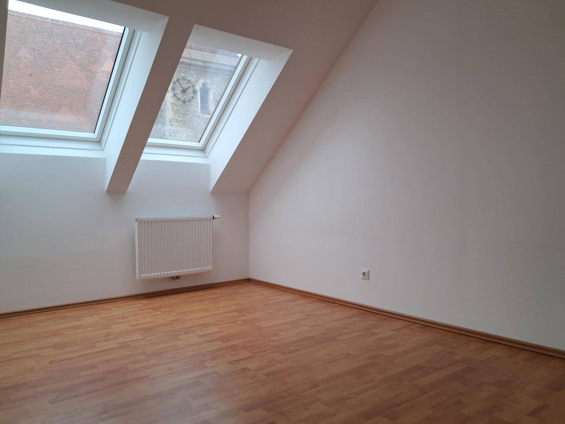 Dachgeschossraum mit zwei Dachfenstern, die einen Blick auf die Dächer der Nachbargebäude ermöglichen.