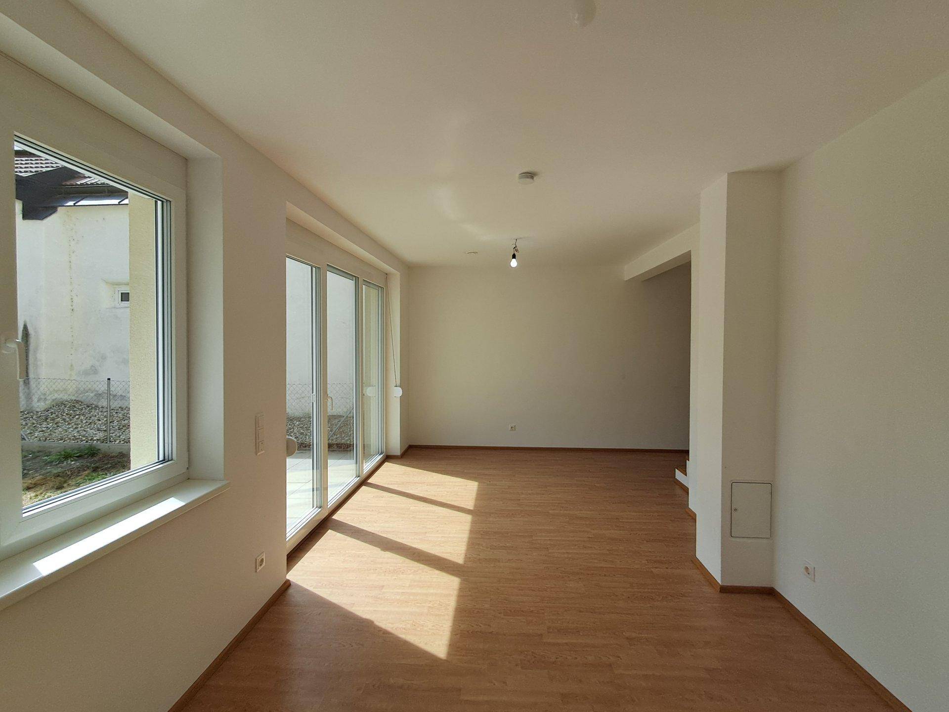 Lichtdurchfluteter Wohnraum mit Laminatboden und großen Fenstern, die Sonnenlicht hereinlassen.