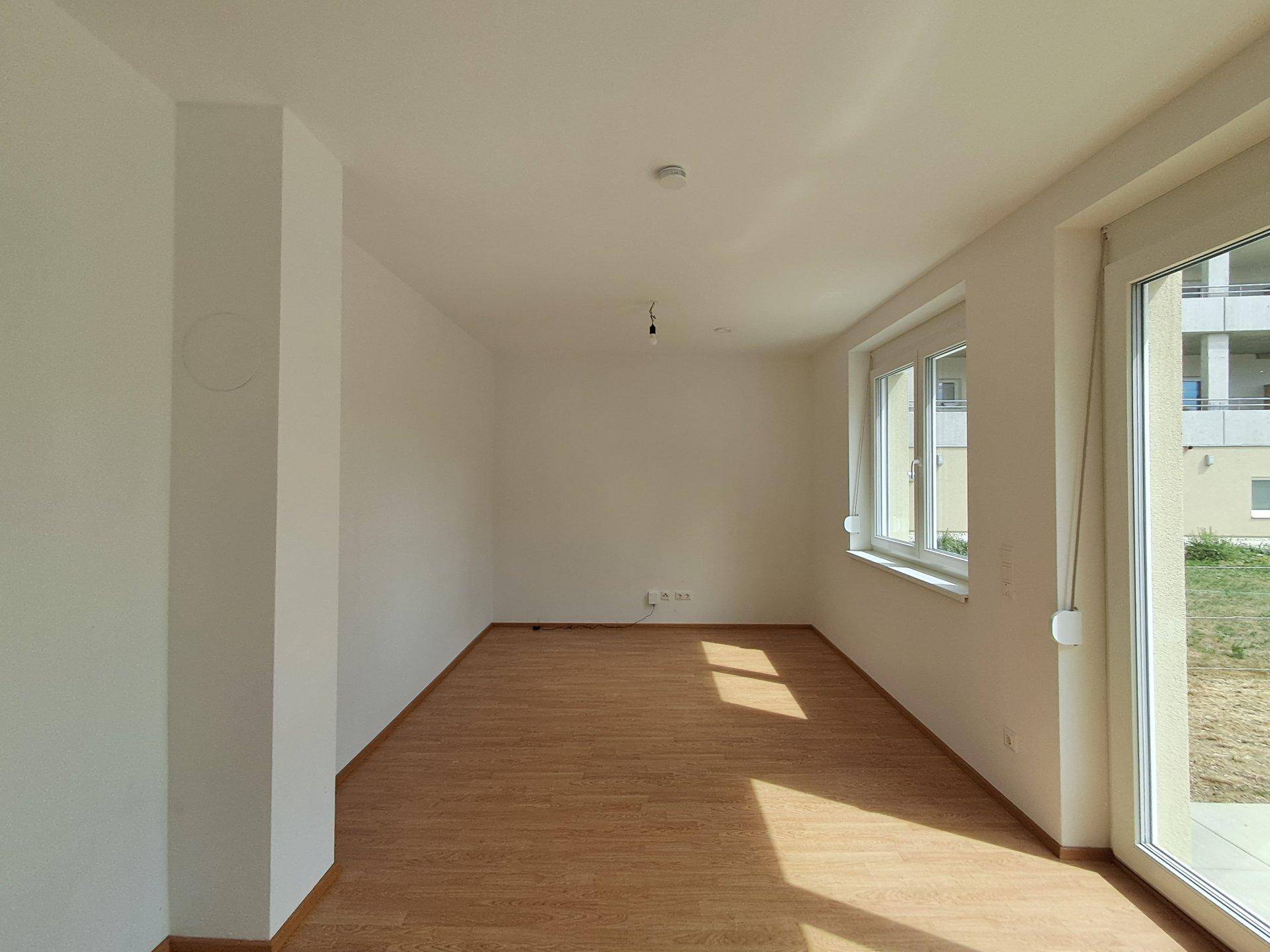 Leeres Wohnzimmer mit Laminatboden und breiten Fenstern, die einen Blick nach draußen bieten.