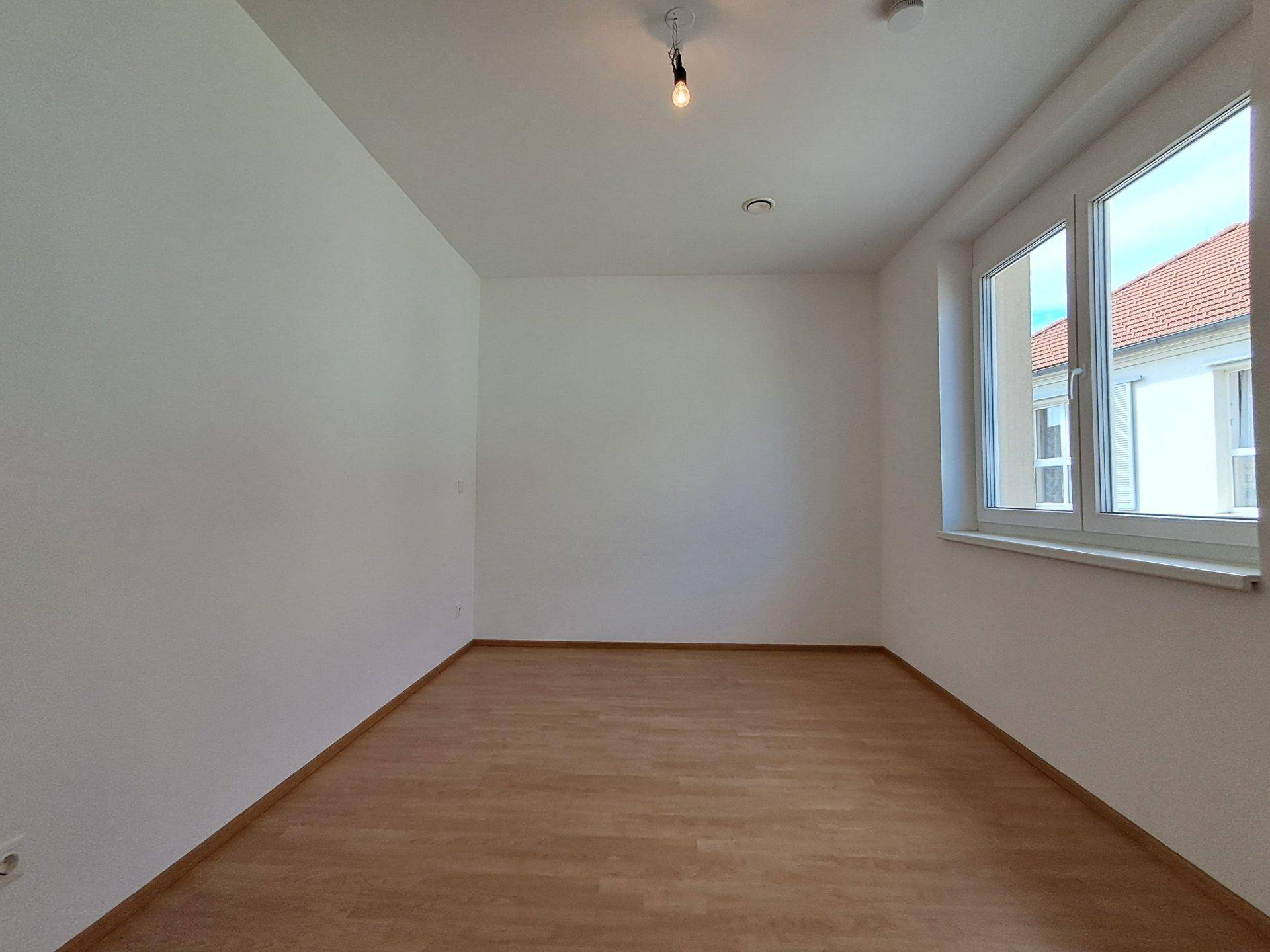 Leeres Schlafzimmer mit Laminatboden und Fenster, das den Raum mit Licht füllt.