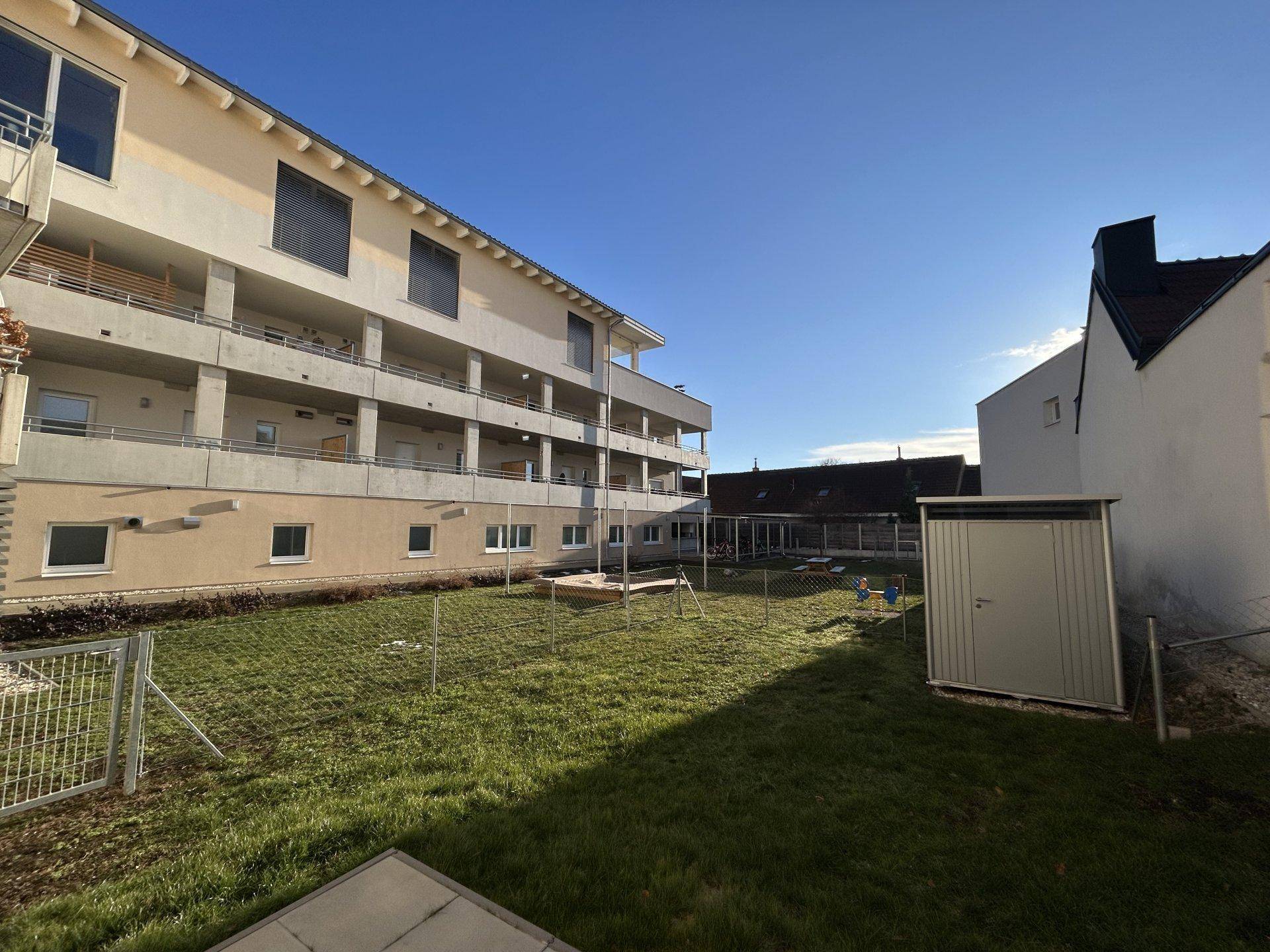 Großer Gartenbereich mit Rasen, Spielplatz und Gartenhaus, umgeben von Wohngebäuden.