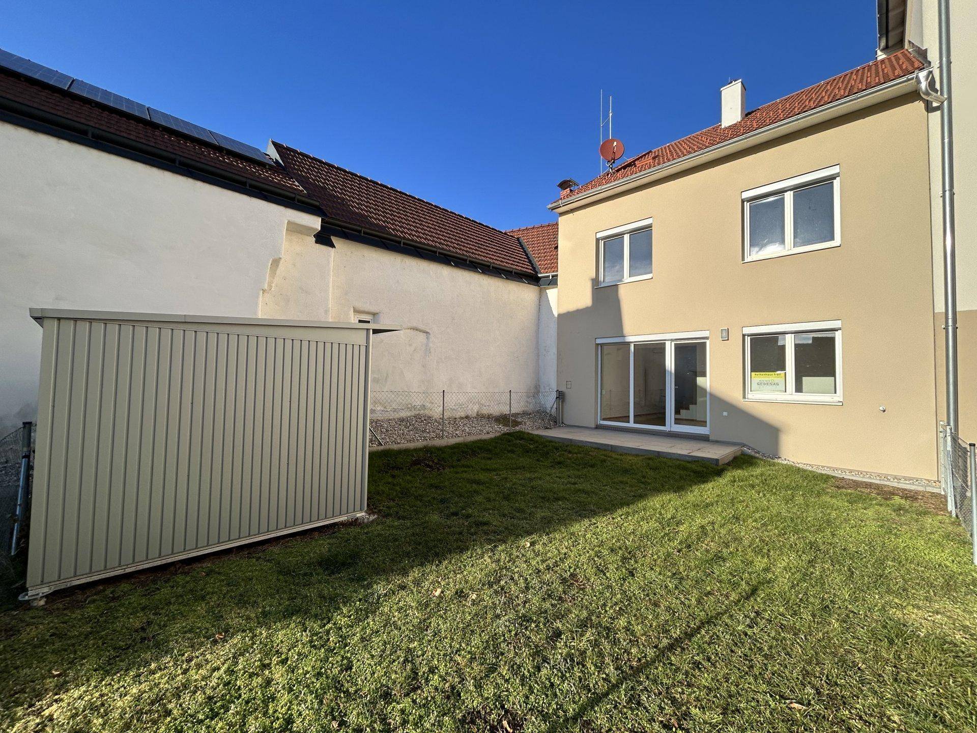 Hinterhof mit Rasenfläche, Gartenhaus und Zugang zu einer Terrasse.