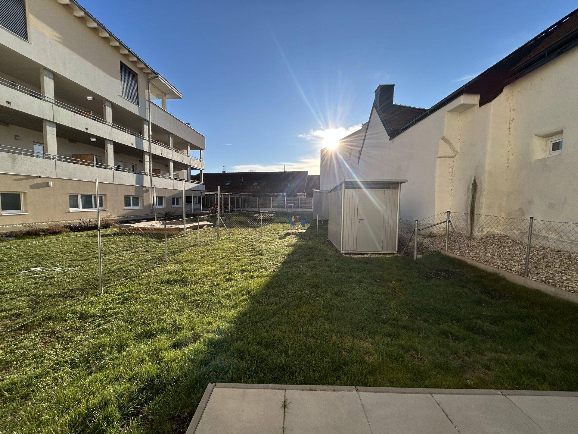 Sonniger Gartenbereich mit Rasenfläche, Gartenhaus und Blick auf umliegende Gebäude.