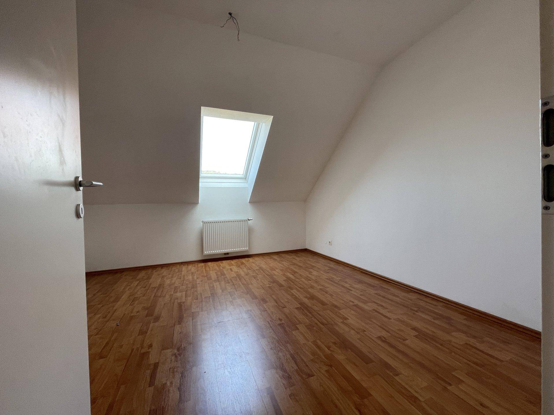 Heller, leerer Raum im Dachgeschoss mit Holzfußboden und einem Dachfenster.