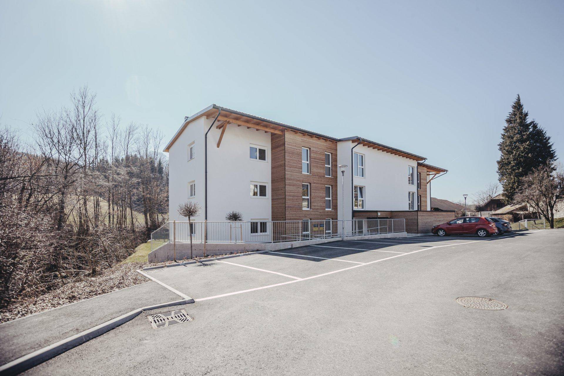 Neubau-Mehrfamilienhaus mit moderner Holzfassade und Parkplätzen in ländlicher Umgebung.