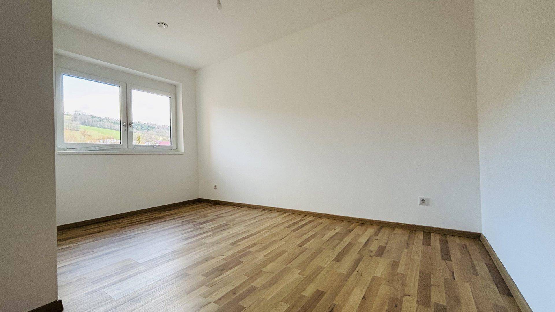 Helles, leeres Zimmer mit Holzparkettboden und Fenster mit Blick ins Grüne.