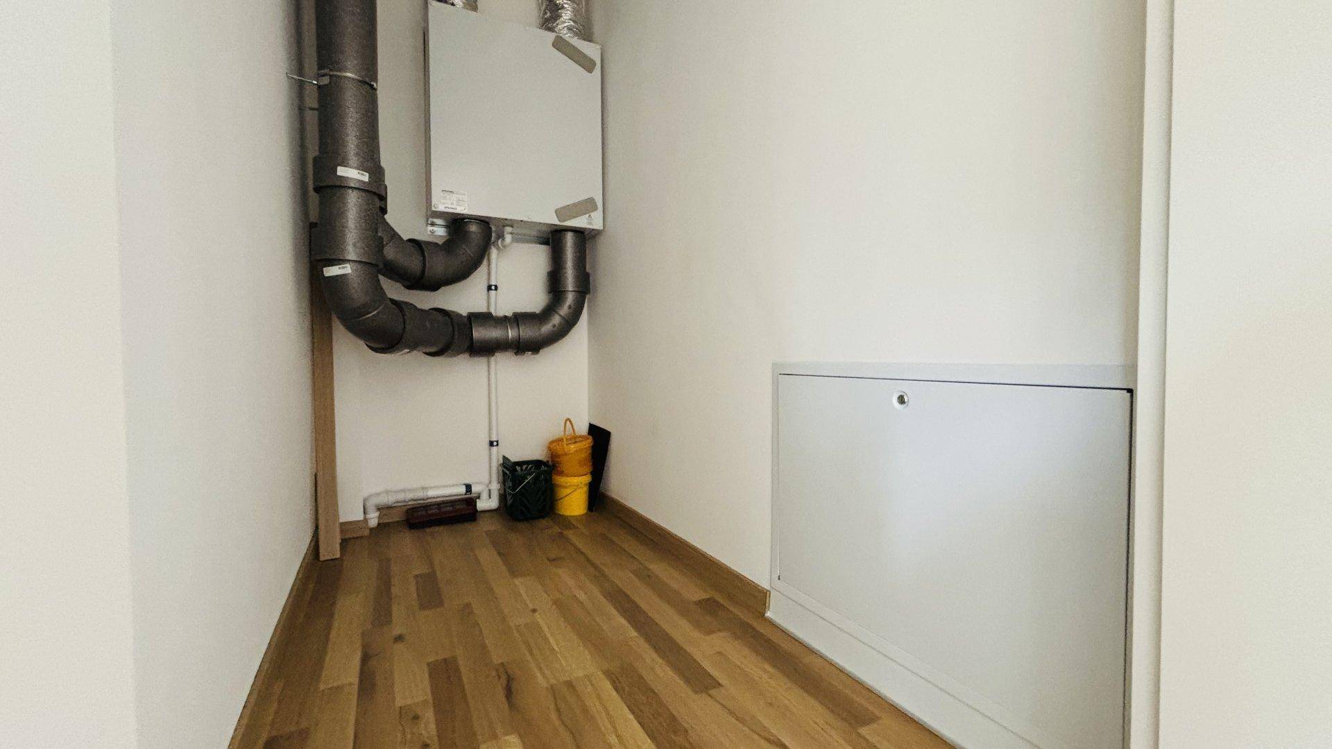Technikraum mit Lüftungsanlage und sichtbaren Rohren, ausgestattet mit Holzboden.