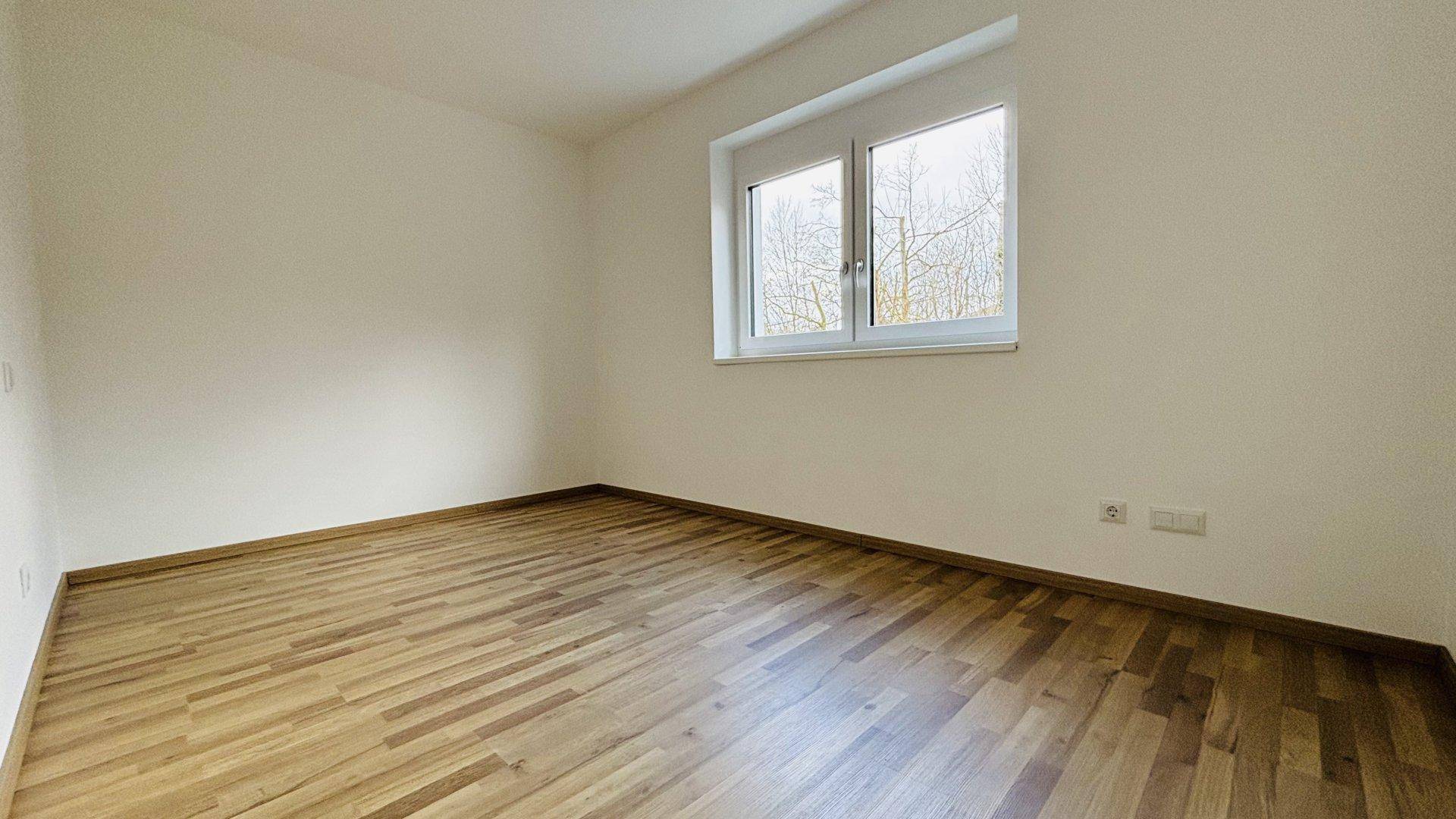 Leeres, helles Zimmer mit Holzparkettboden und Fenster mit Blick ins Grüne.