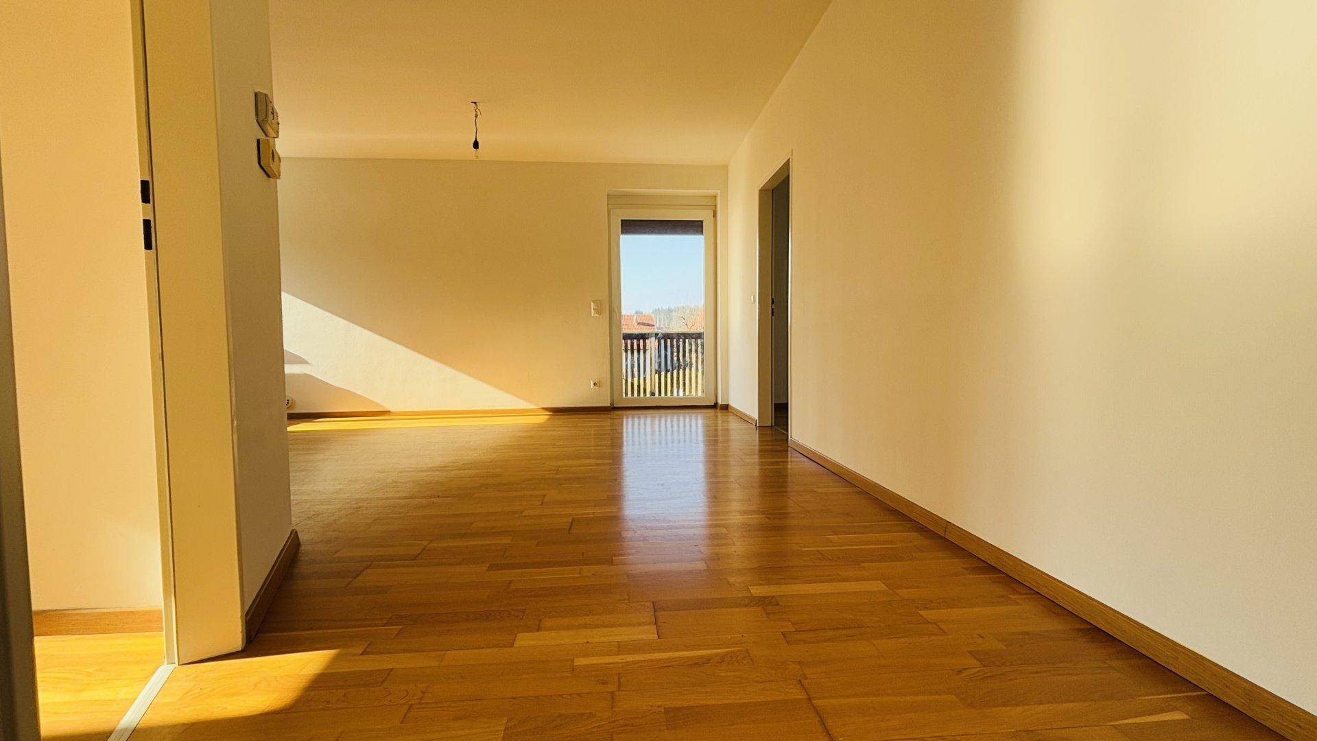 Helles, leeres Zimmer mit Holzboden und Zugang zu einem Balkon, durchflutet von Sonnenlicht.