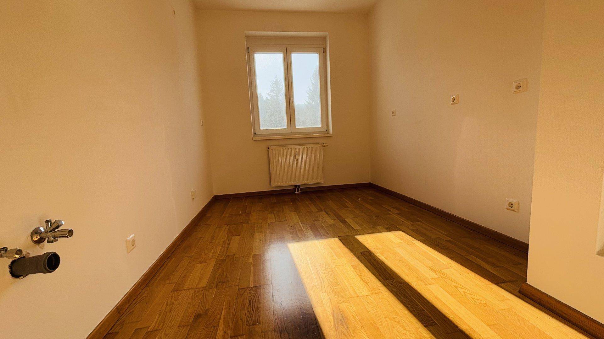 Leerer Raum mit Holzboden und Fenster, ausgestattet mit Anschlüssen für eine Waschmaschine oder Spüle.