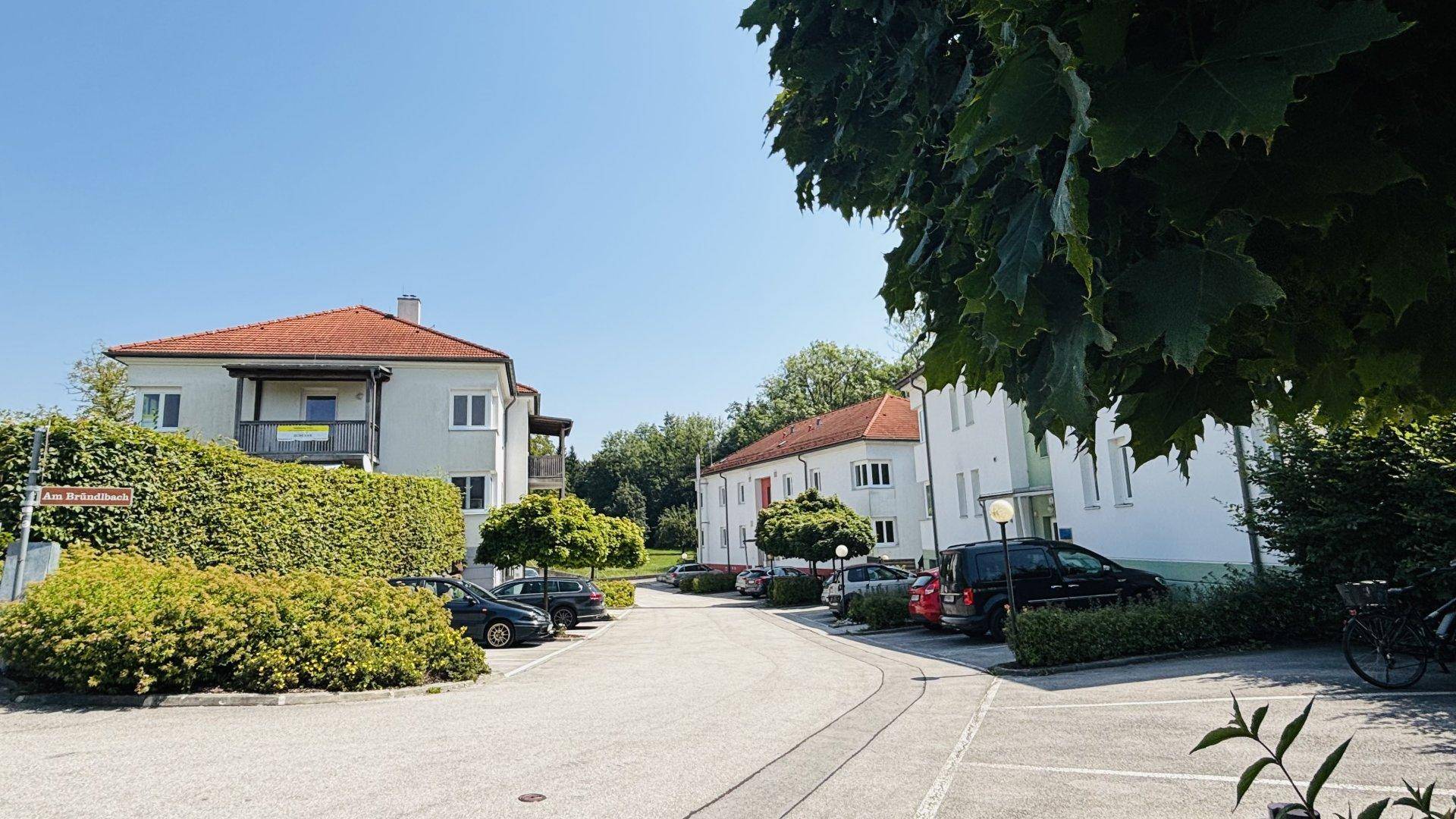 Ruhige Wohnstraße mit parkenden Autos und weiteren Gebäuden, umgeben von viel Grün.