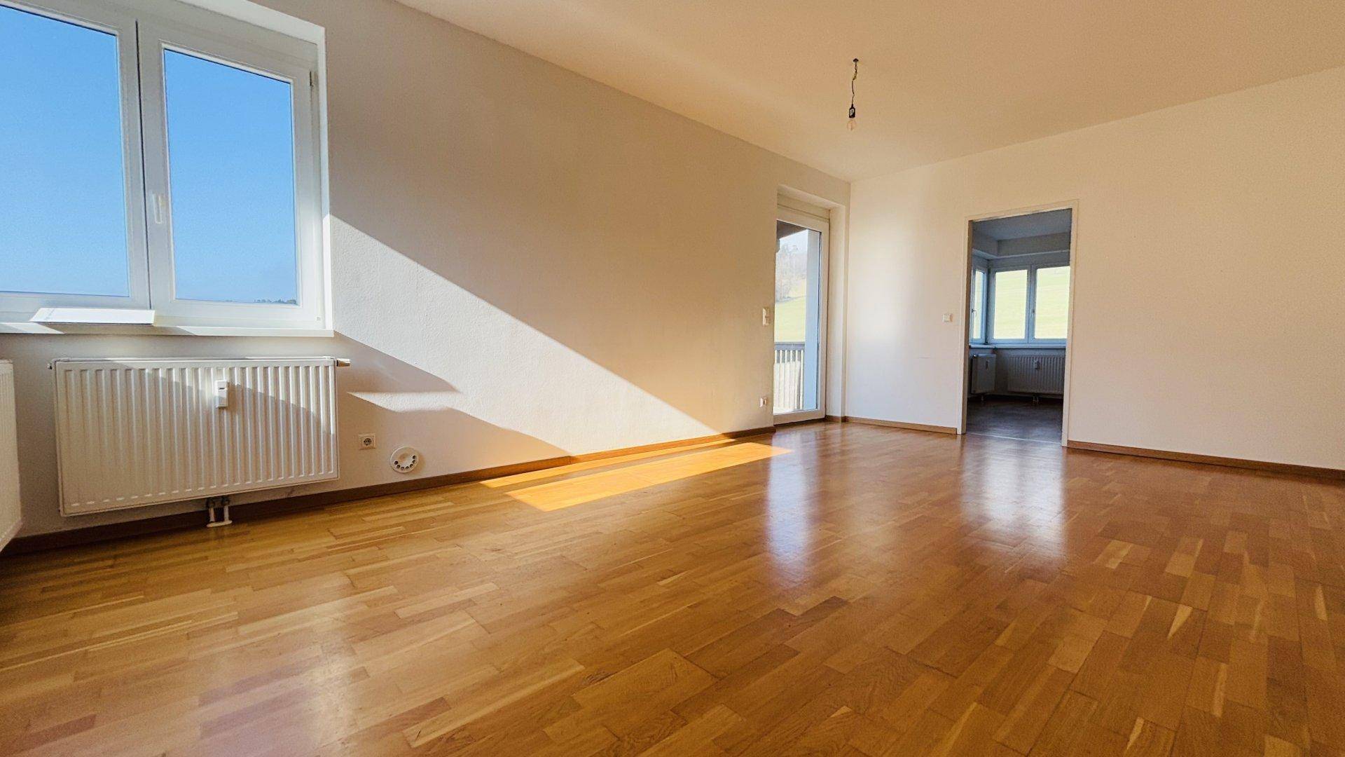 Heller Wohnbereich mit Holzboden, Fenster und Balkonzugang, der zu einem weiteren Zimmer führt.