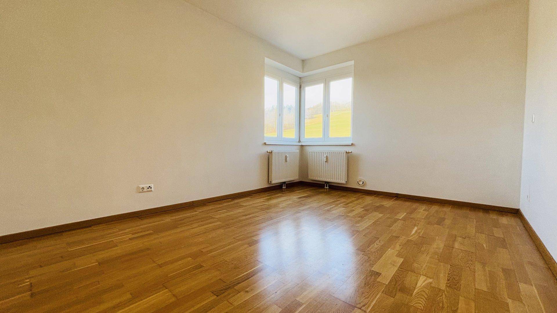 Geräumiges Schlafzimmer mit Holzboden und einem hellen Eckfenster, sonnendurchflutet.