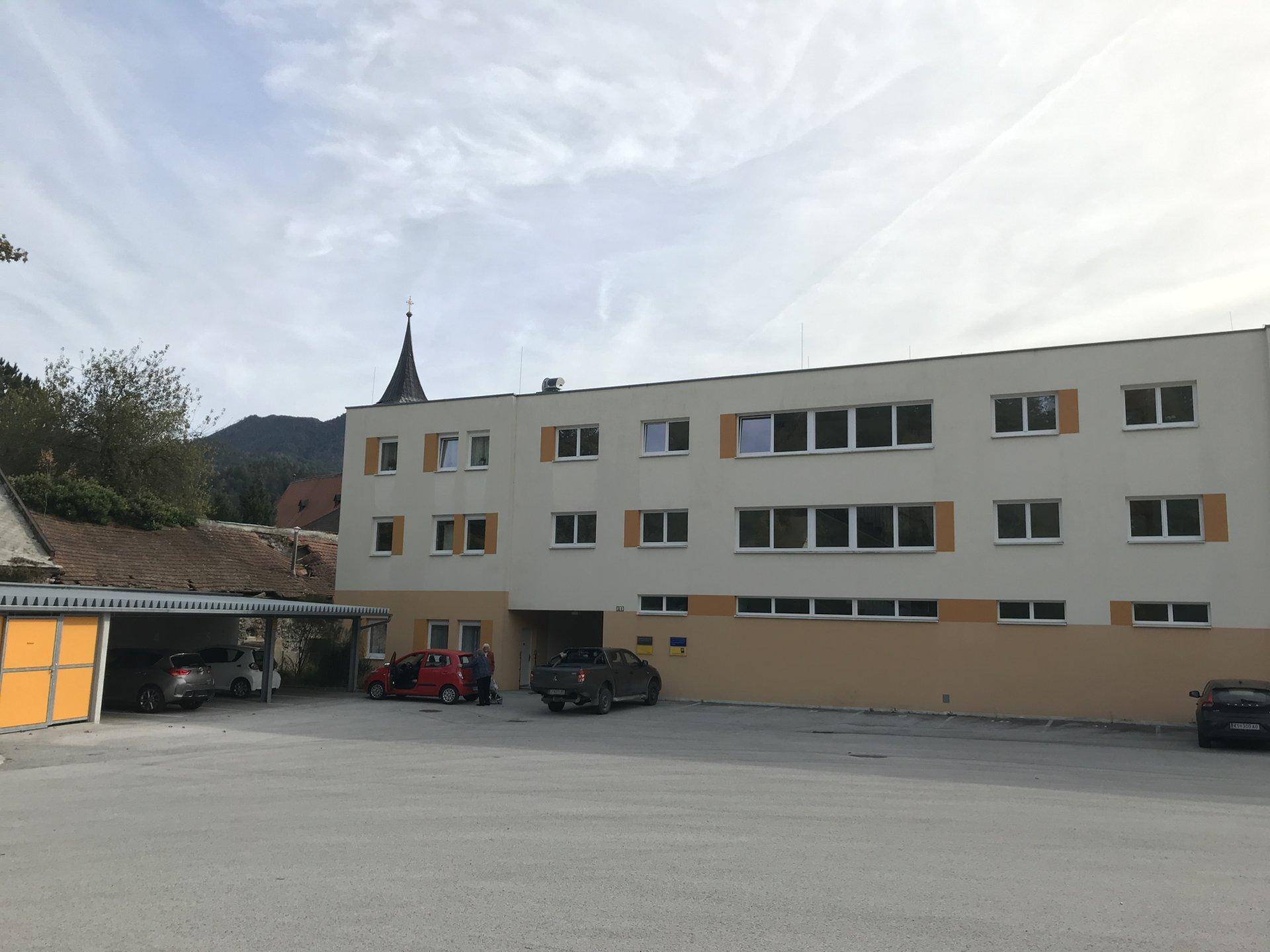 Ansicht eines größeren Gebäudes mit Parkplatz und einem Kirchturm im Hintergrund unter bewölktem Himmel.