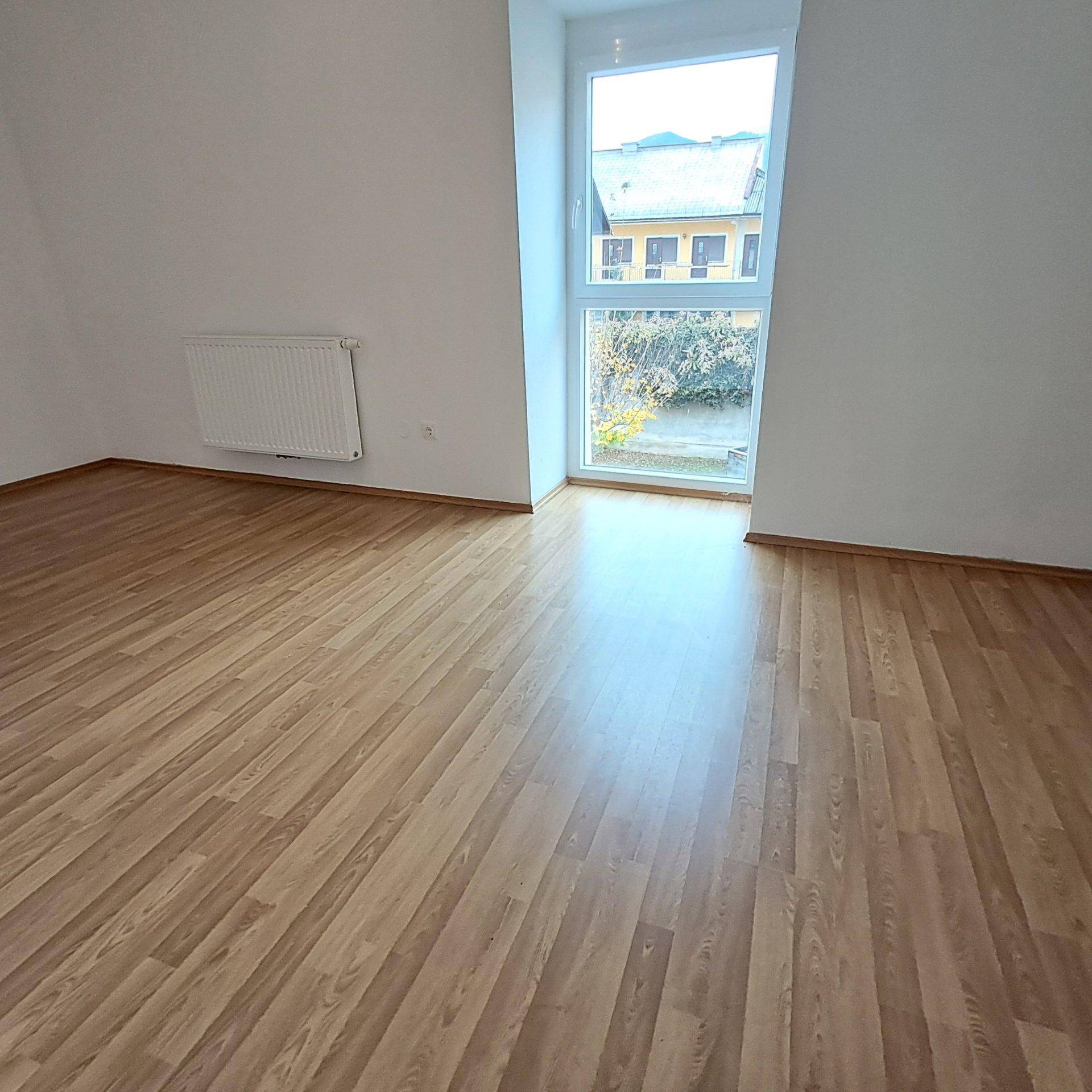 Unmöblierter Raum mit hellem Laminatboden und einem großen Fenster, das viel Tageslicht hereinlässt.