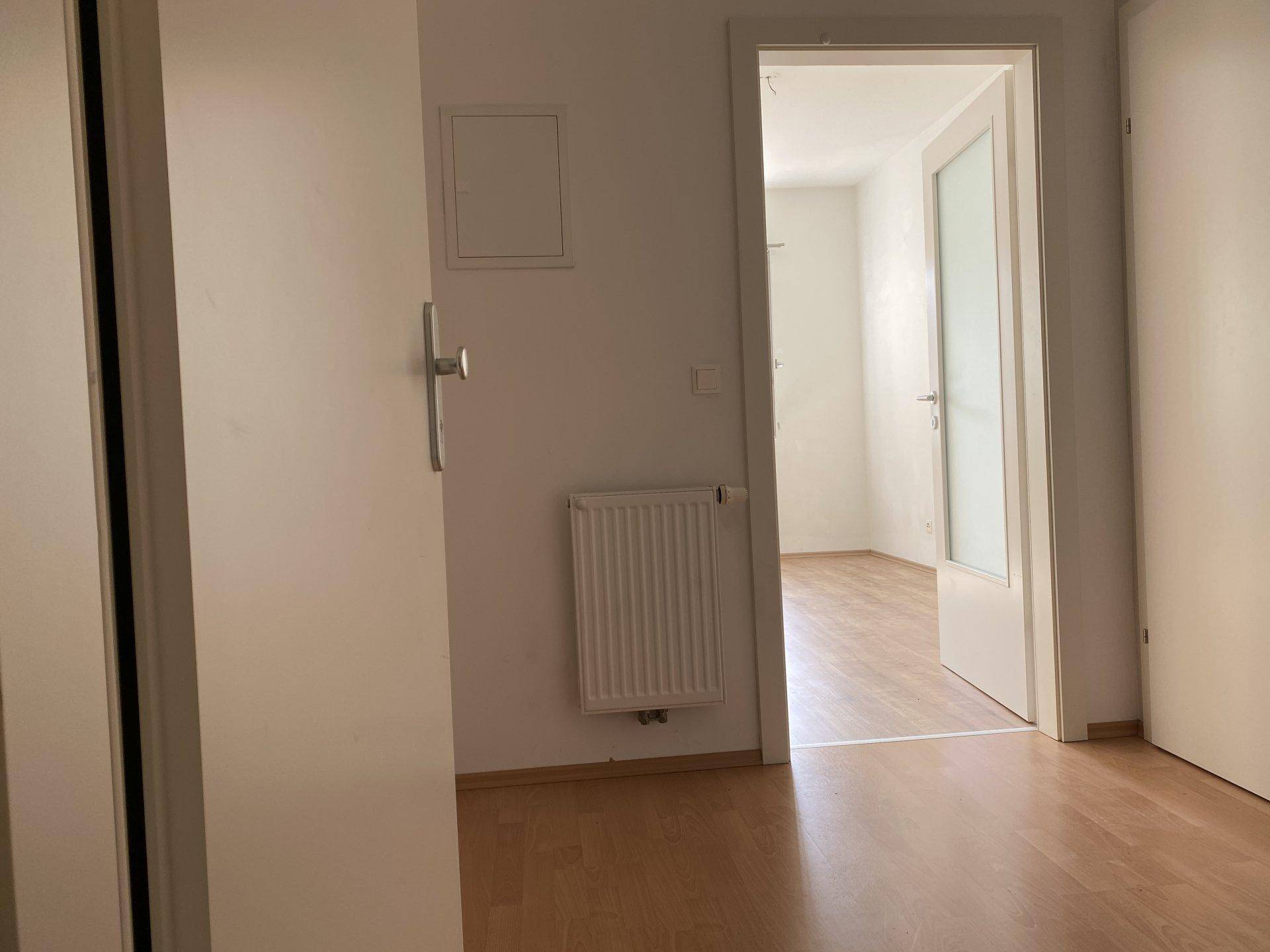 Heller Flur mit Heizkörper und Zugang zu einem weiteren leeren Zimmer mit Holzboden.