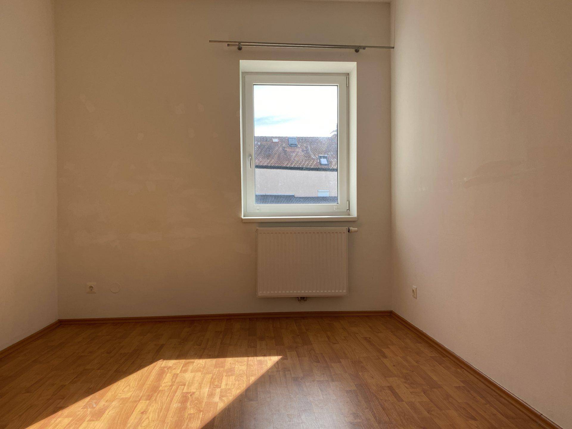 Heller, leerer Raum mit einem Fenster und Laminatboden, der viel Sonnenlicht hereinlässt.
