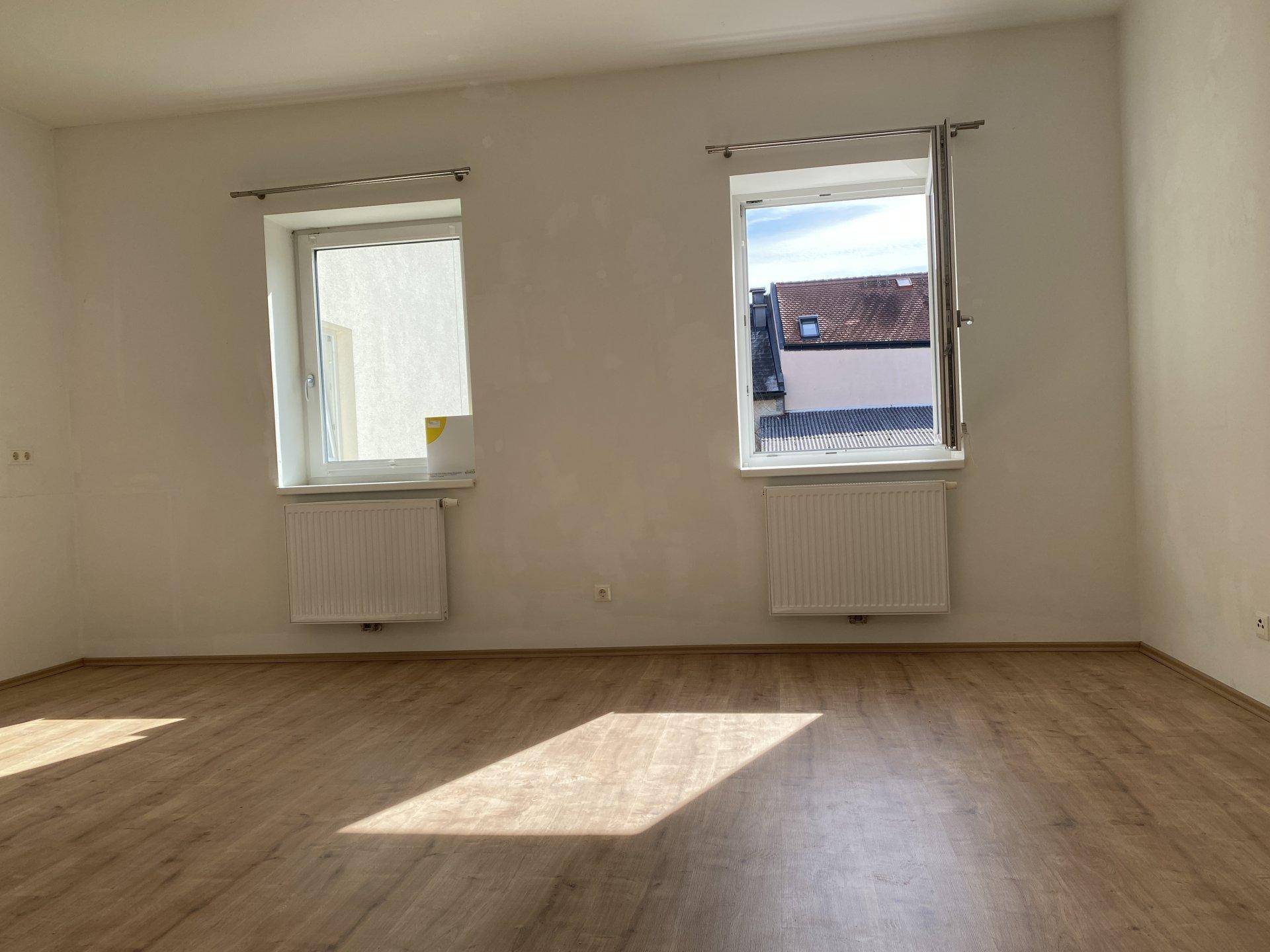 Leerer Raum mit zwei Fenstern, eines davon geöffnet, und hellem Laminatboden, gut belichtet.