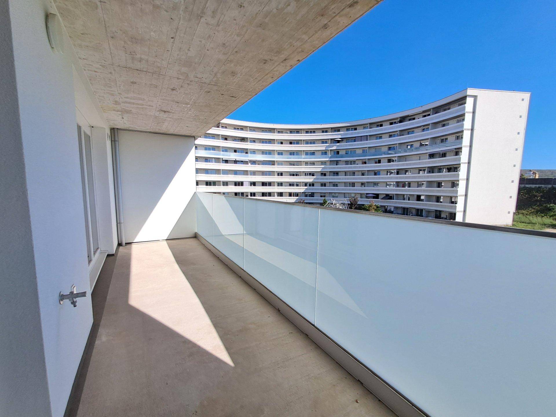 Geräumiger Balkon mit Glasbrüstung und Blick auf ein gegenüberliegendes modernes Gebäude.