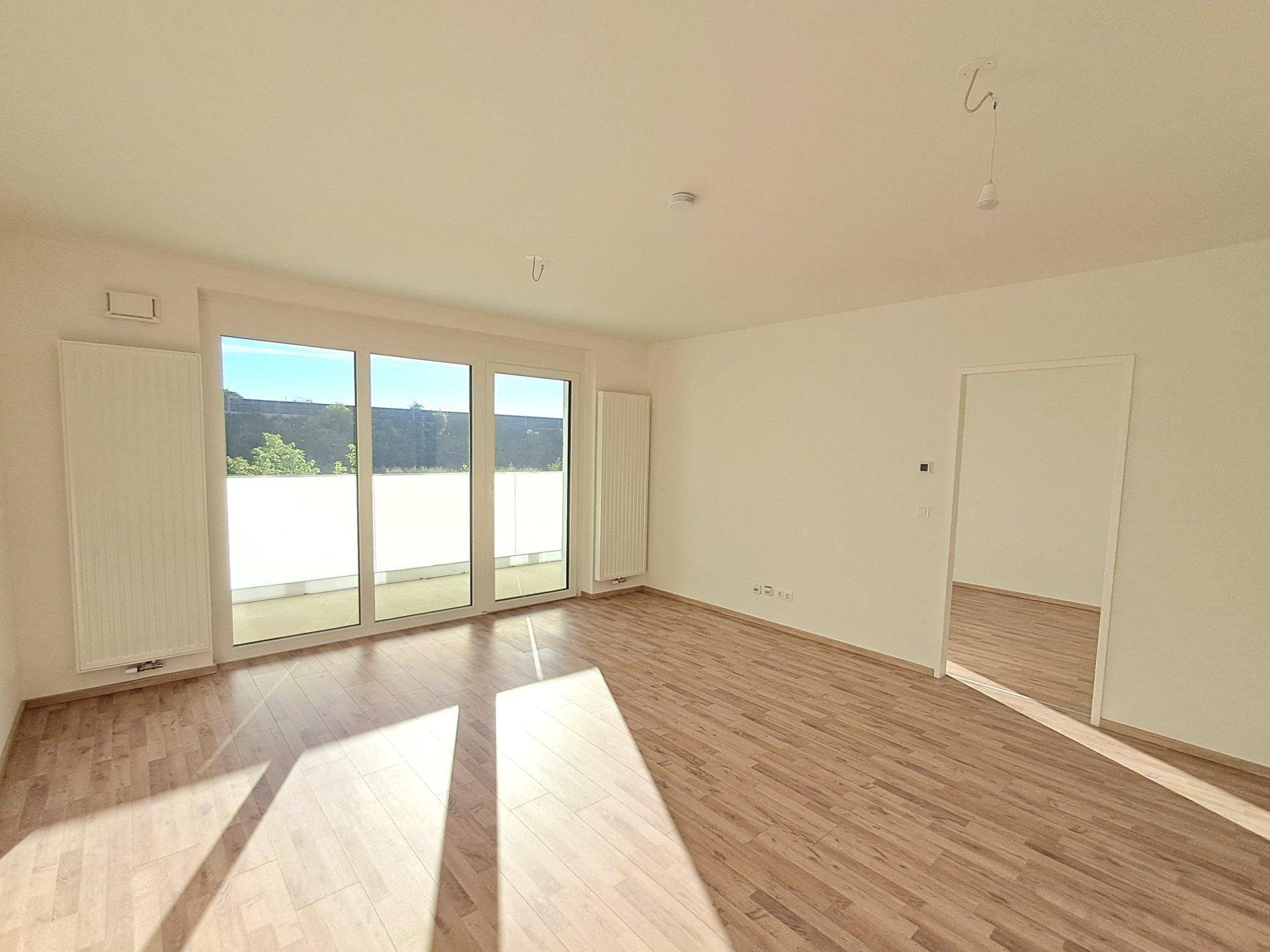 Geräumiger Wohnraum mit hellem Holzfußboden und viel Tageslicht durch große Fenster.